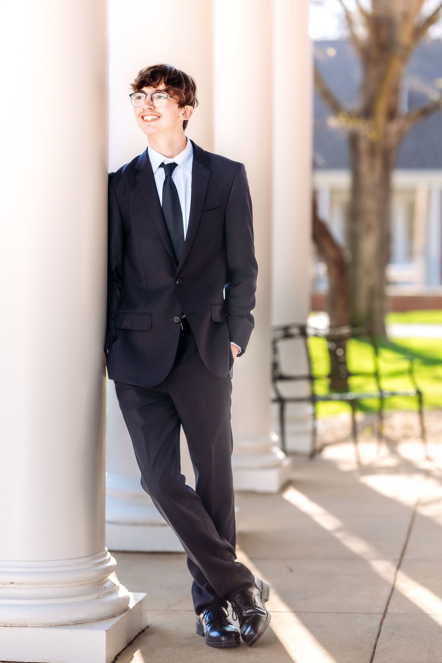 A person in a black suit and tie leans casually against a white column on a porch with trees in the background.