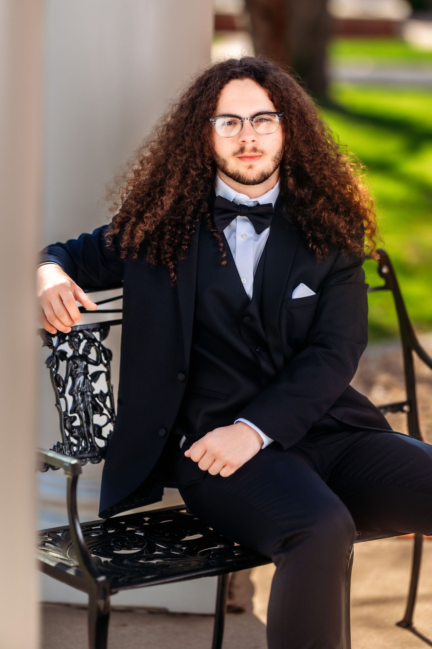 Person with long curly hair wearing formal black suit poses outdoors on bench for graduation photos.
