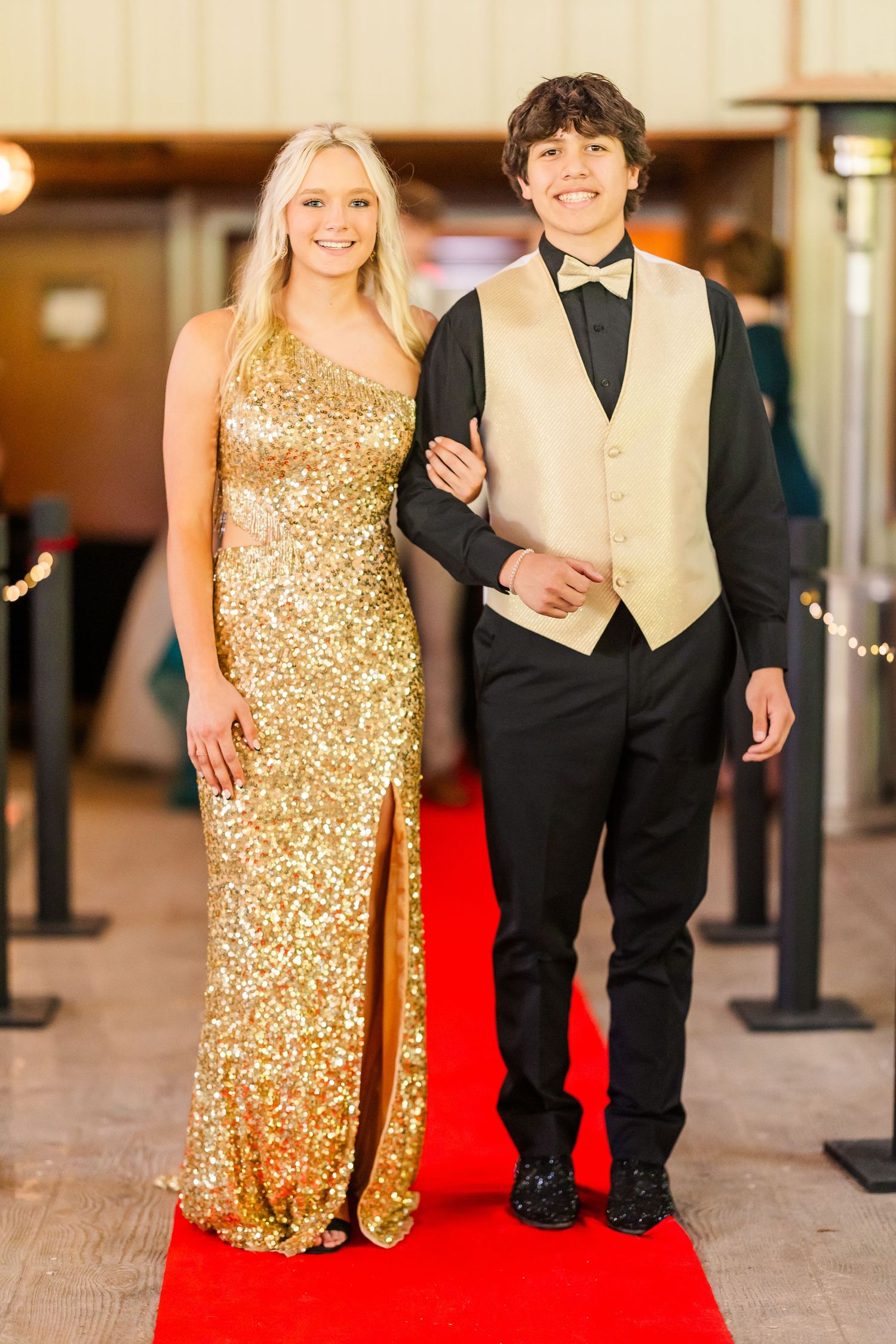 Couples walking the red carpet in gold sequined dresses and formal suits at an evening event.