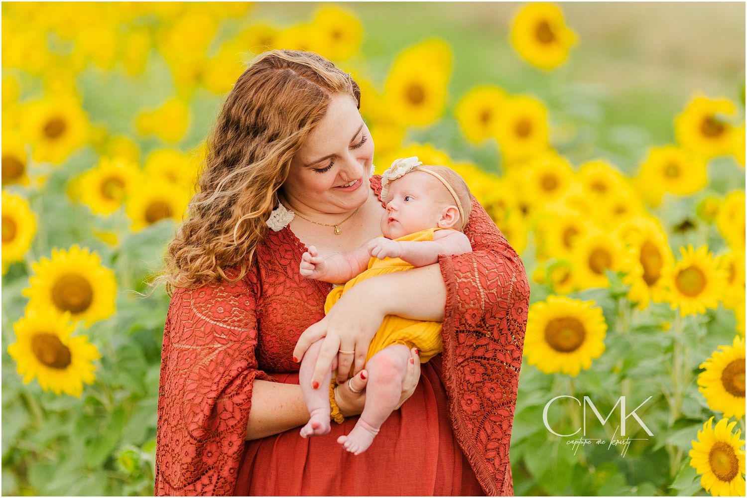 A tender moment captured between parent and infant in a vibrant sunflower field wearing rust-colored clothing.