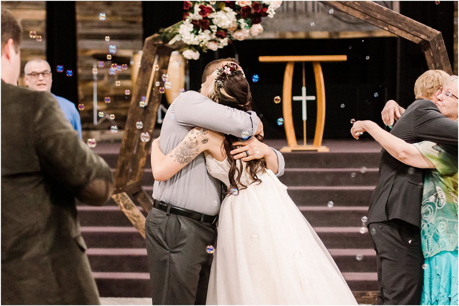Wedding guests embrace the bride in her white gown under a rustic wooden arch with floral decor.
