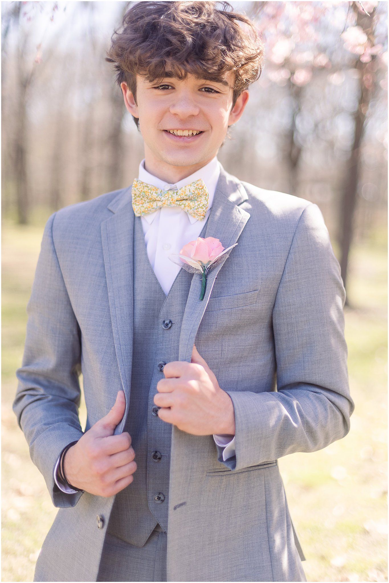 Person wearing grey three piece suit with yellow bowtie and pink boutonniere in spring outdoor setting.