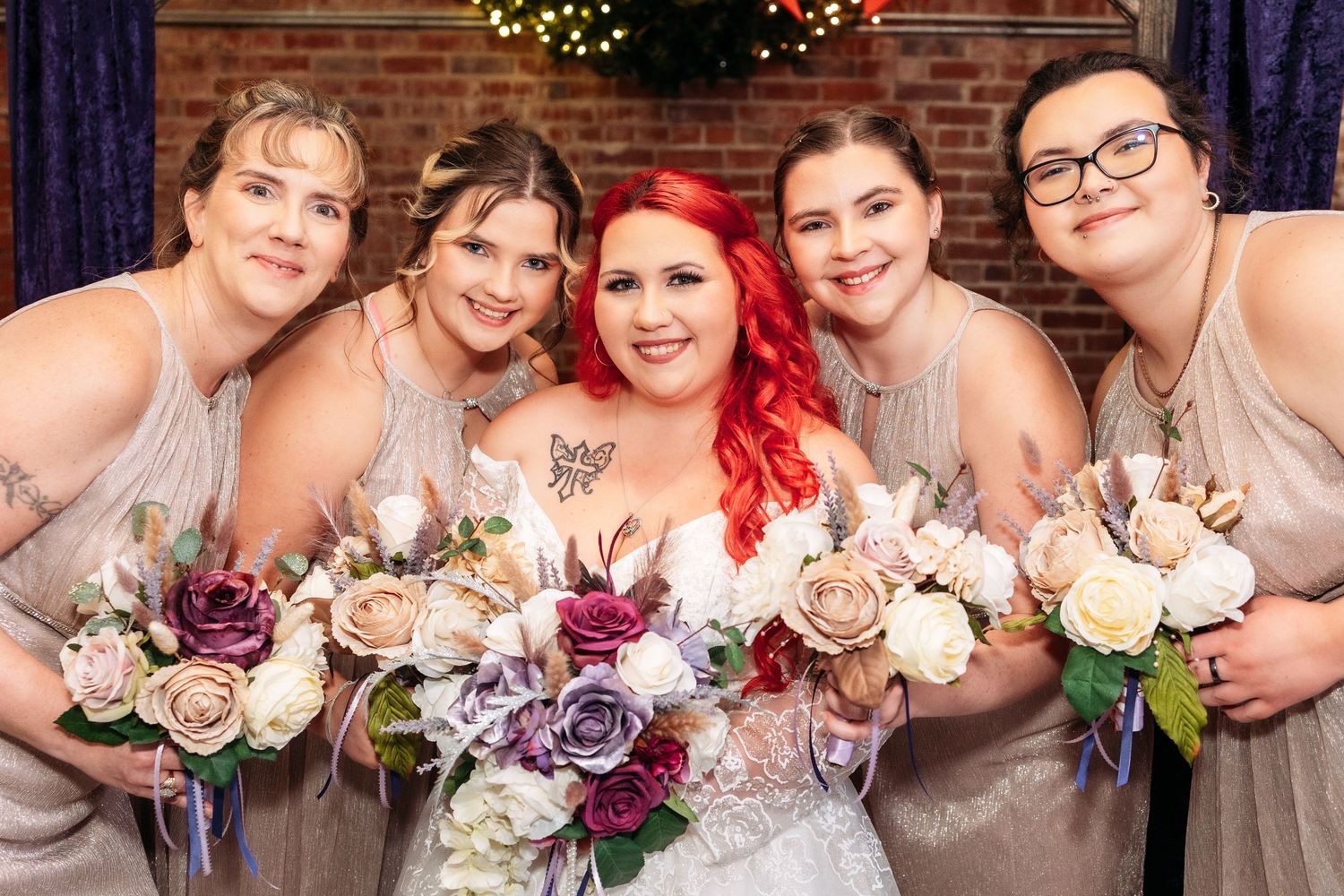 Bridal party poses with bouquets in neutral dresses against a brick wall backdrop.