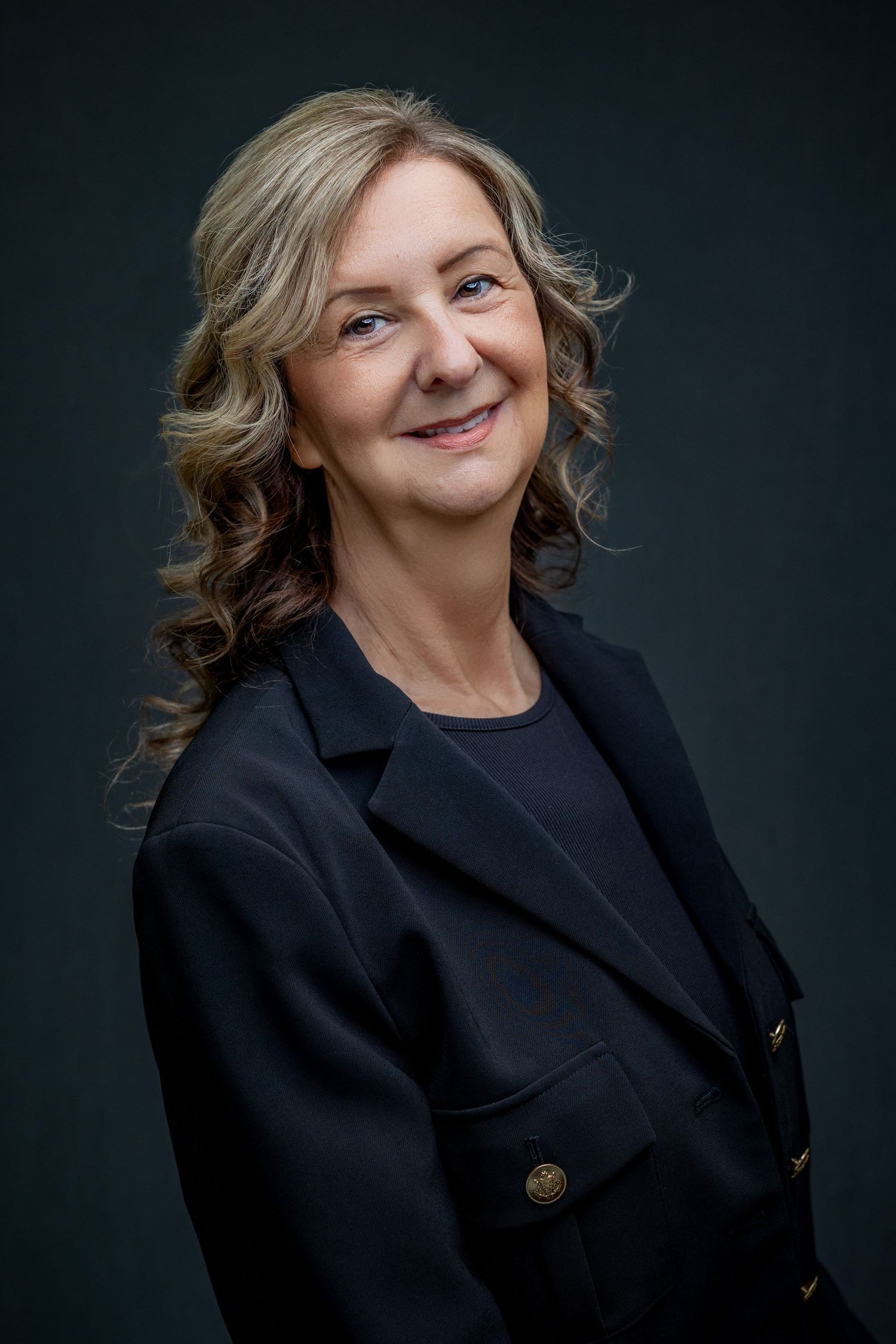 Professional portrait of woman in black blazer with wavy hair against dark background.