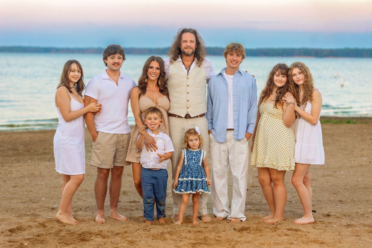 Large family group portrait on sandy beach at sunset, smiling adults and children in casual summer outfits.