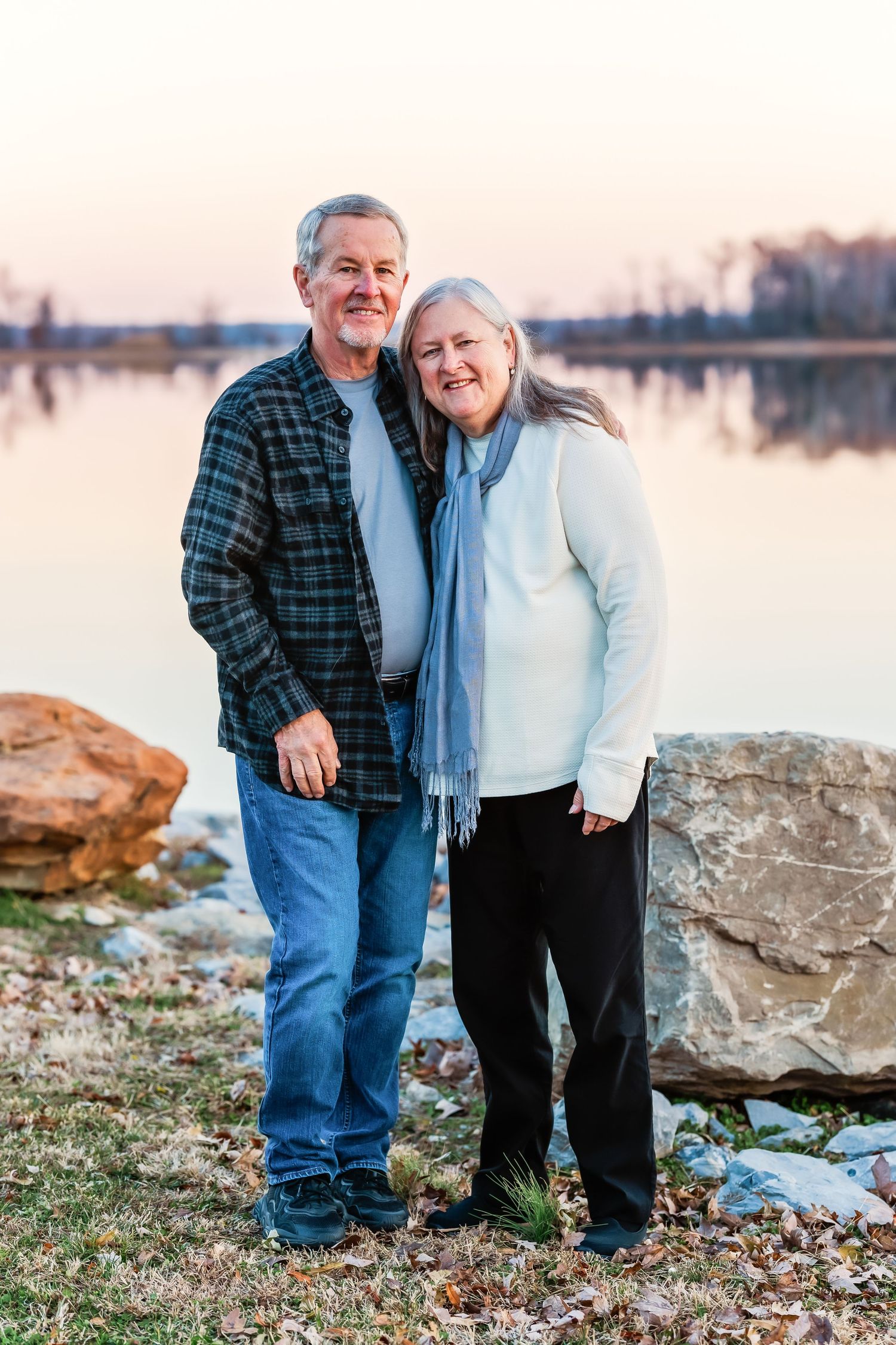 Couple poses together by a lakeside with rocks and fallen leaves during sunset.