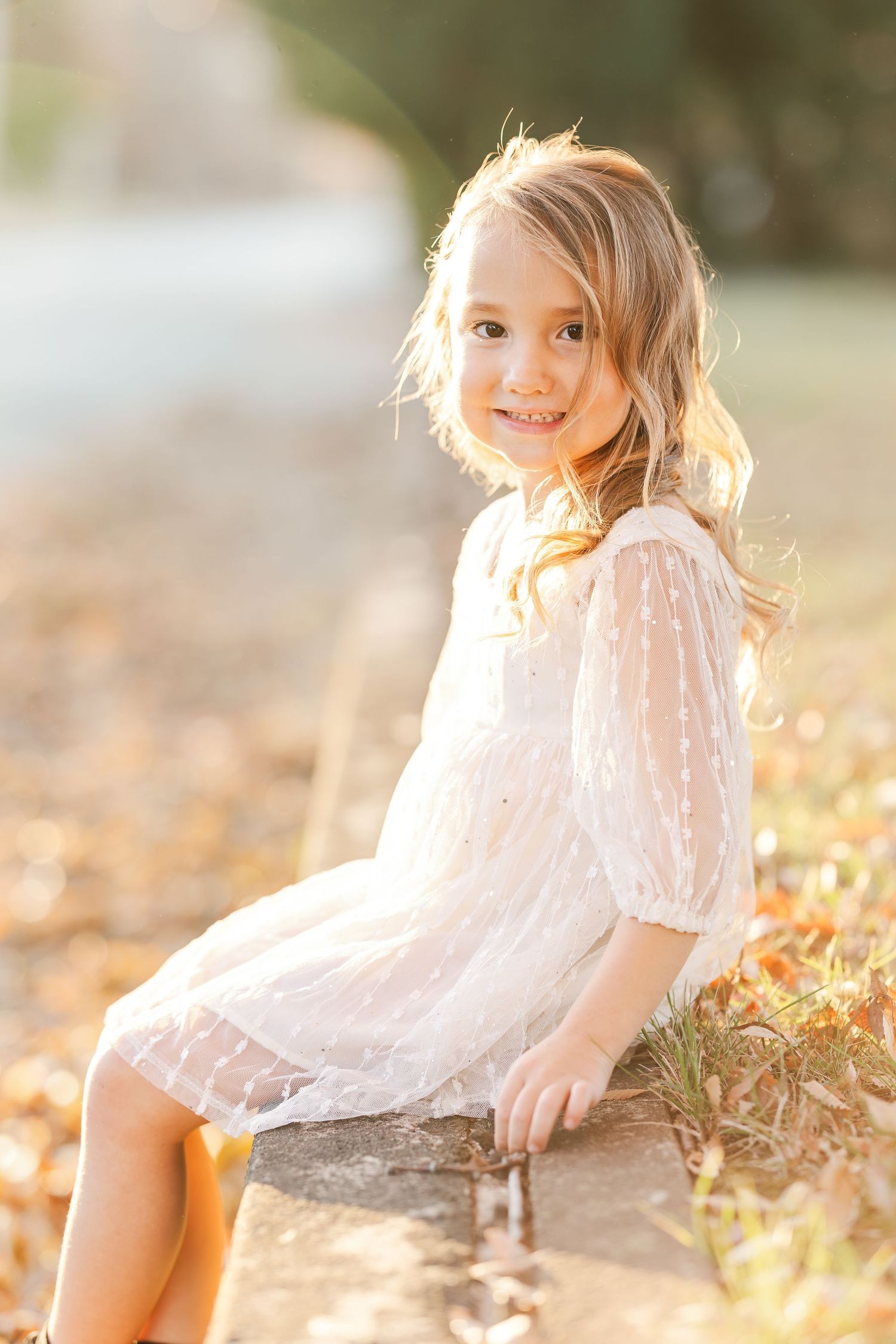 Young girl in white lace dress poses on ground during golden hour.