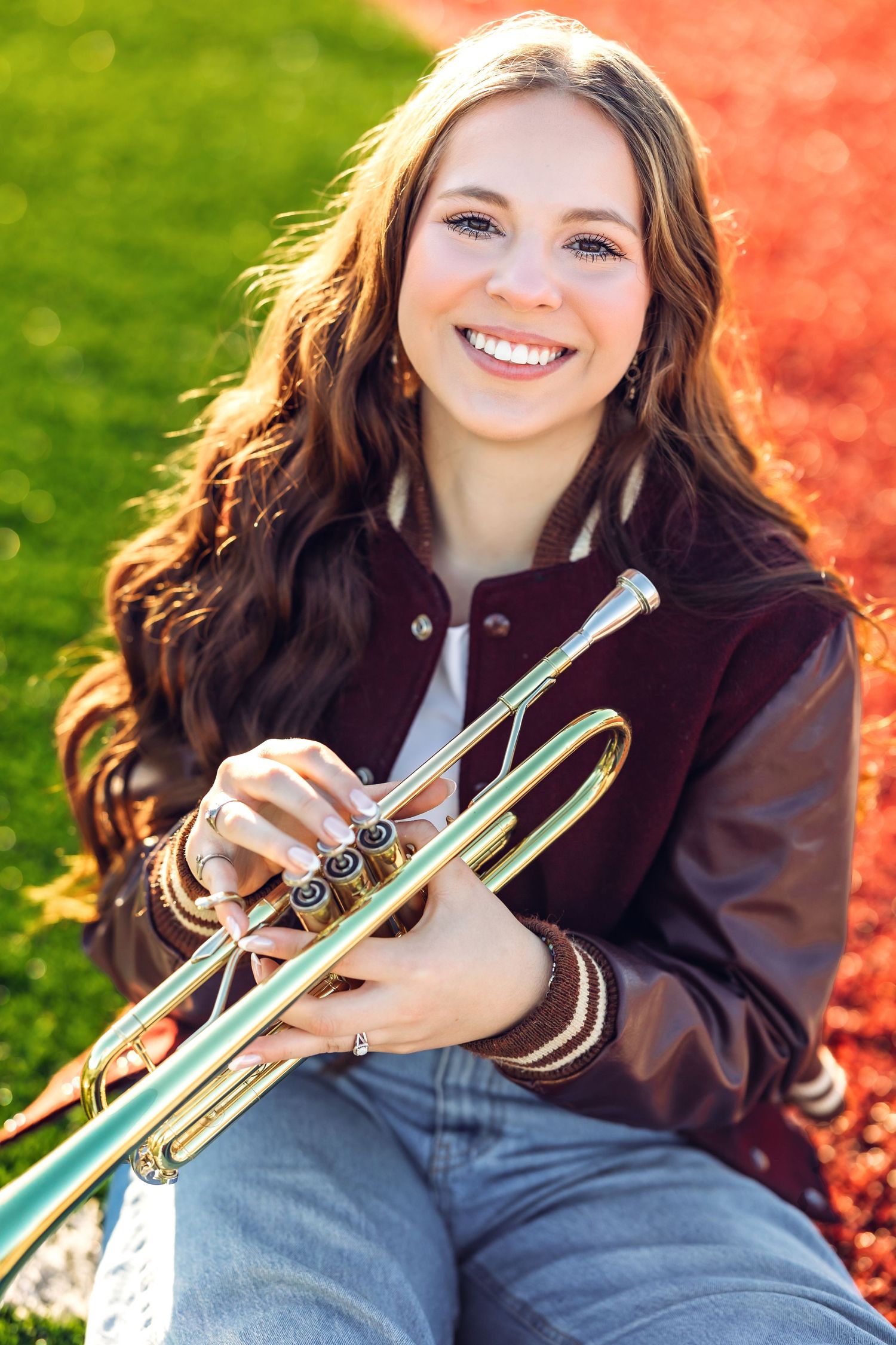 A musician poses with a trumpet while wearing a burgundy jacket against a colorful outdoor backdrop.