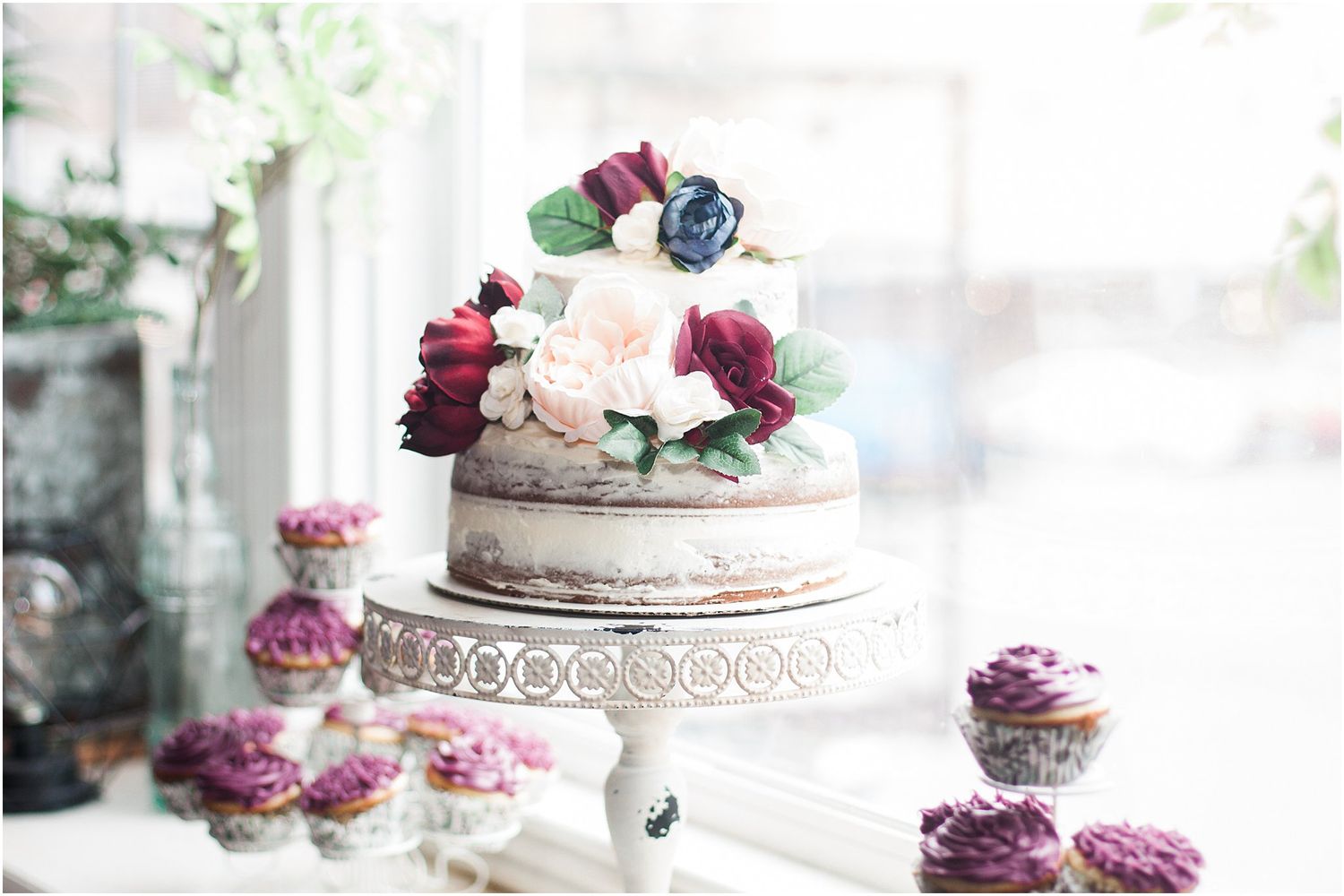 Elegant naked cake with burgundy and pink flowers displayed on an ornate white cake stand.