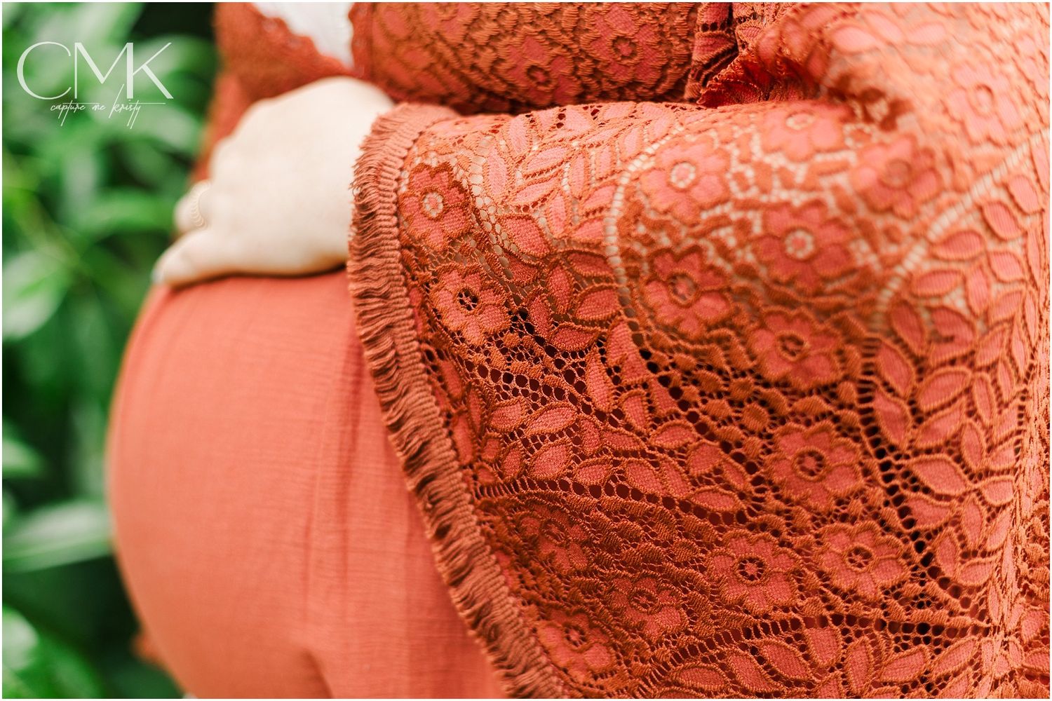 Close-up detail of coral-colored lace maternity dress fabric against green foliage background.