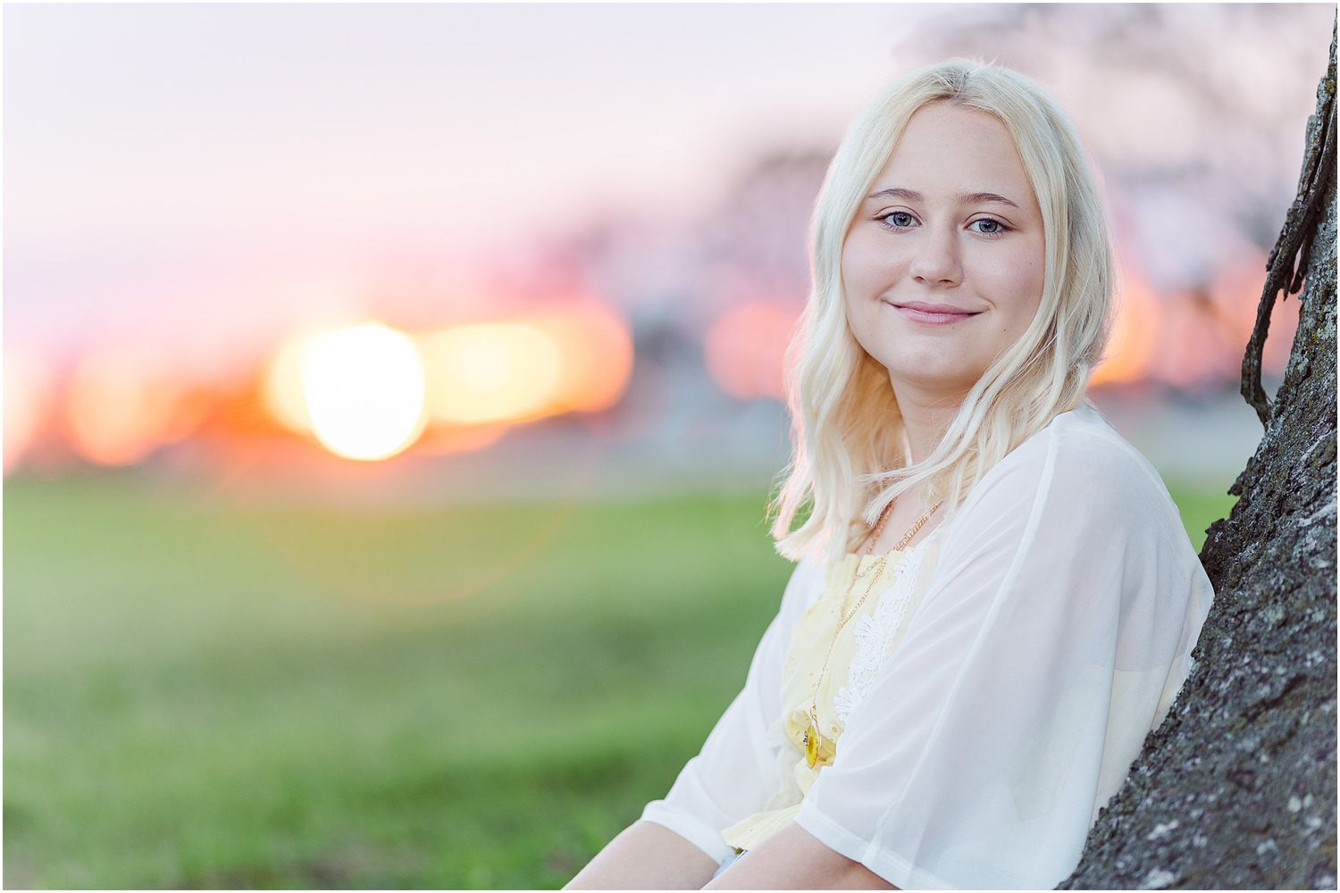 Senior girl leaning against a tree by a corn field with a pink and orange sunset in the background