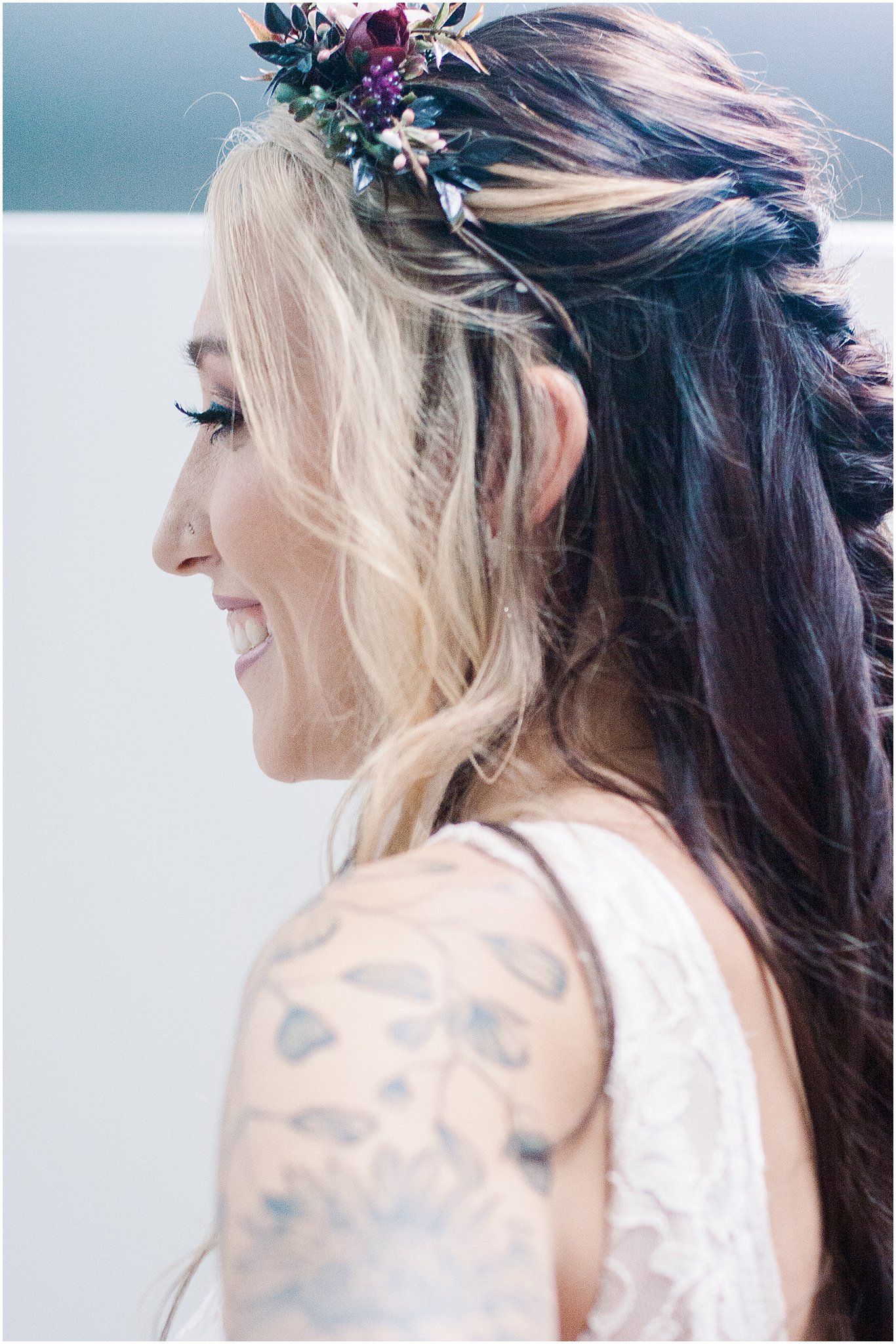 Bride with blonde and dark hair wearing floral crown and white dress shows artistic arm tattoo in profile view.