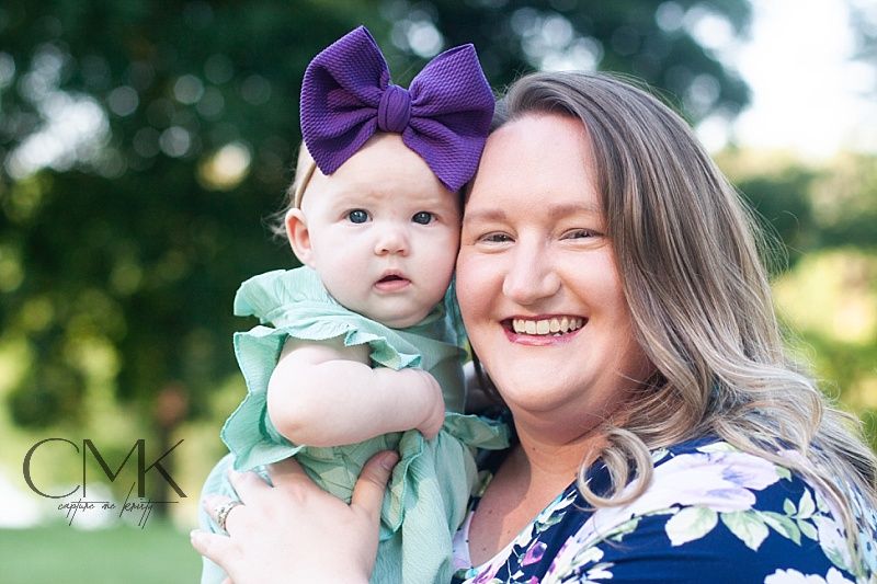 A woman in a navy floral dress holds a baby wearing mint green and purple bow outdoors.