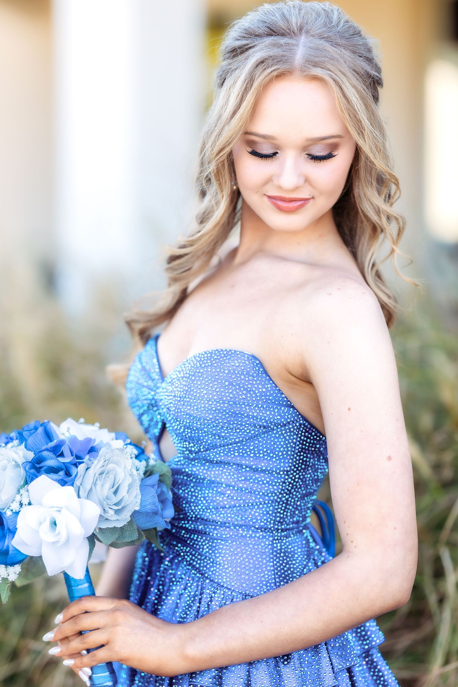 Woman in sparkly blue strapless dress holds matching blue bouquet in outdoor setting.