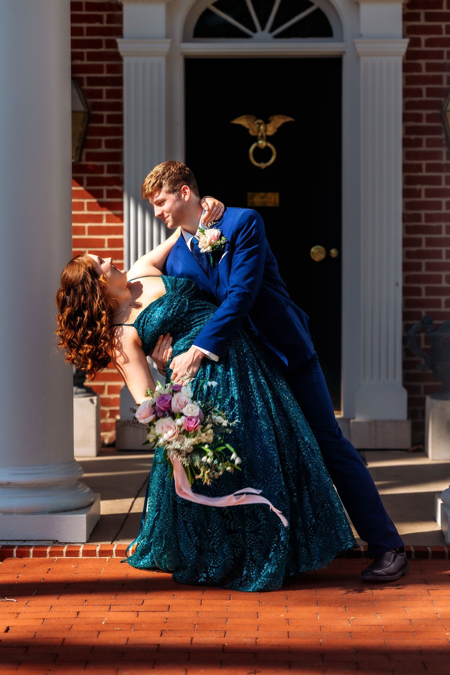 A romantic couple in formal attire share an elegant dip pose in front of a brick building with a black door and white columns.