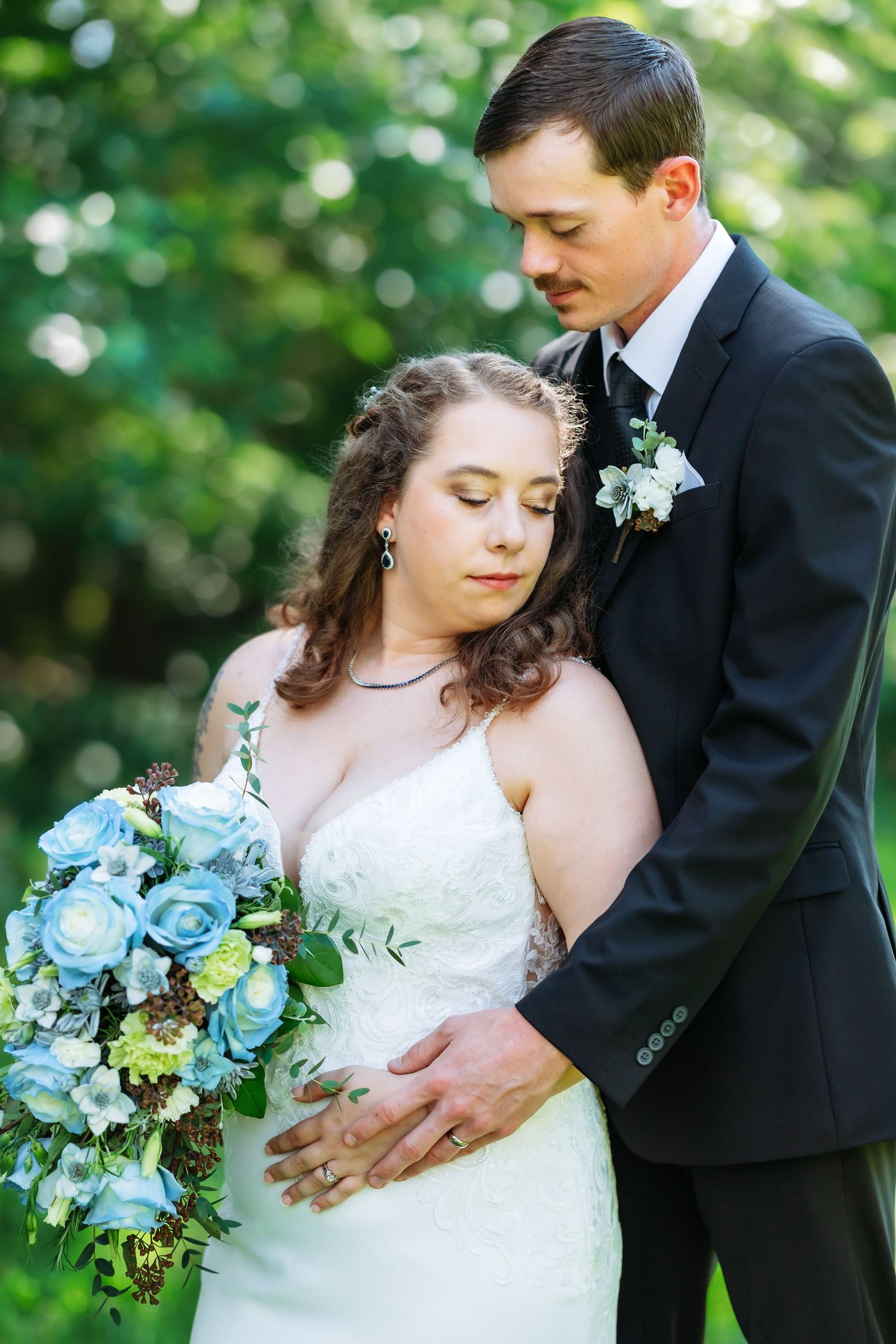 Elegant wedding couple embracing in a sunlit garden setting with blue floral bouquet.