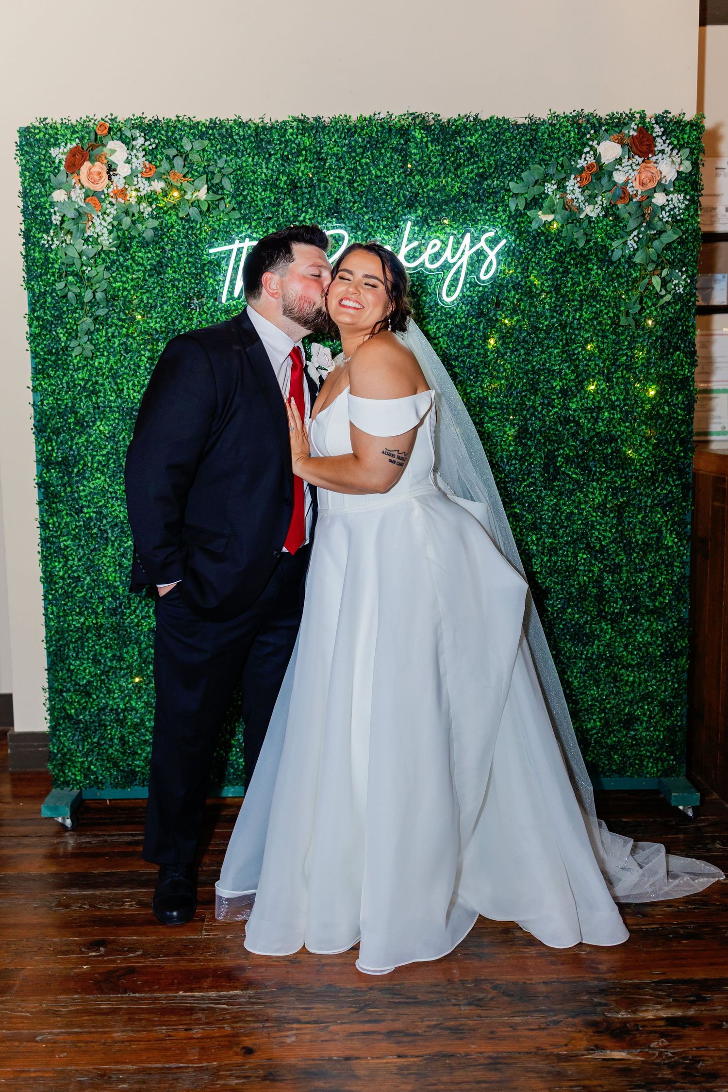 Wedding couple posing in front of a green floral wall backdrop with neon signage.