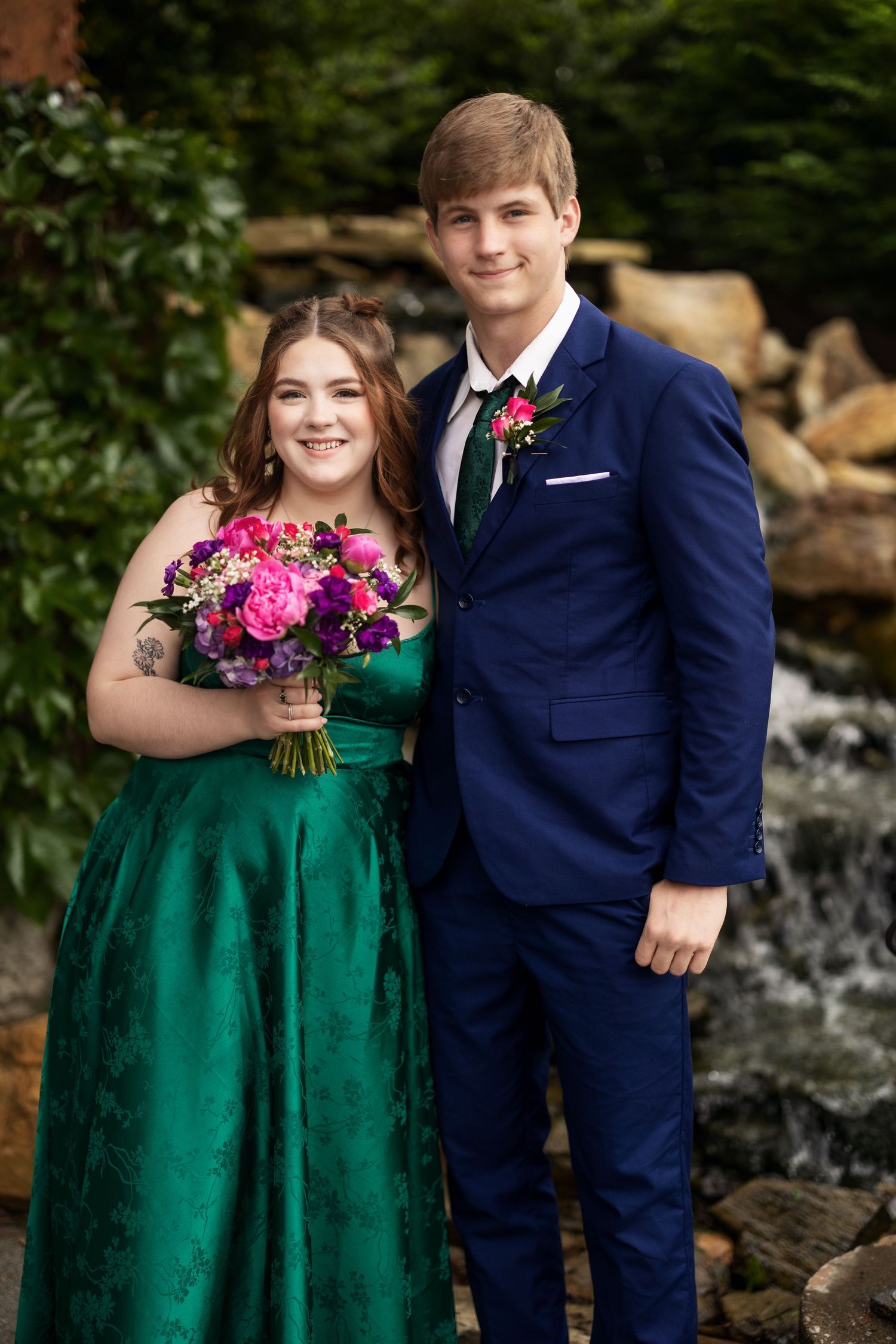 A couple poses in prom attire, she in emerald gown with bouquet, he in navy suit with boutonniere.