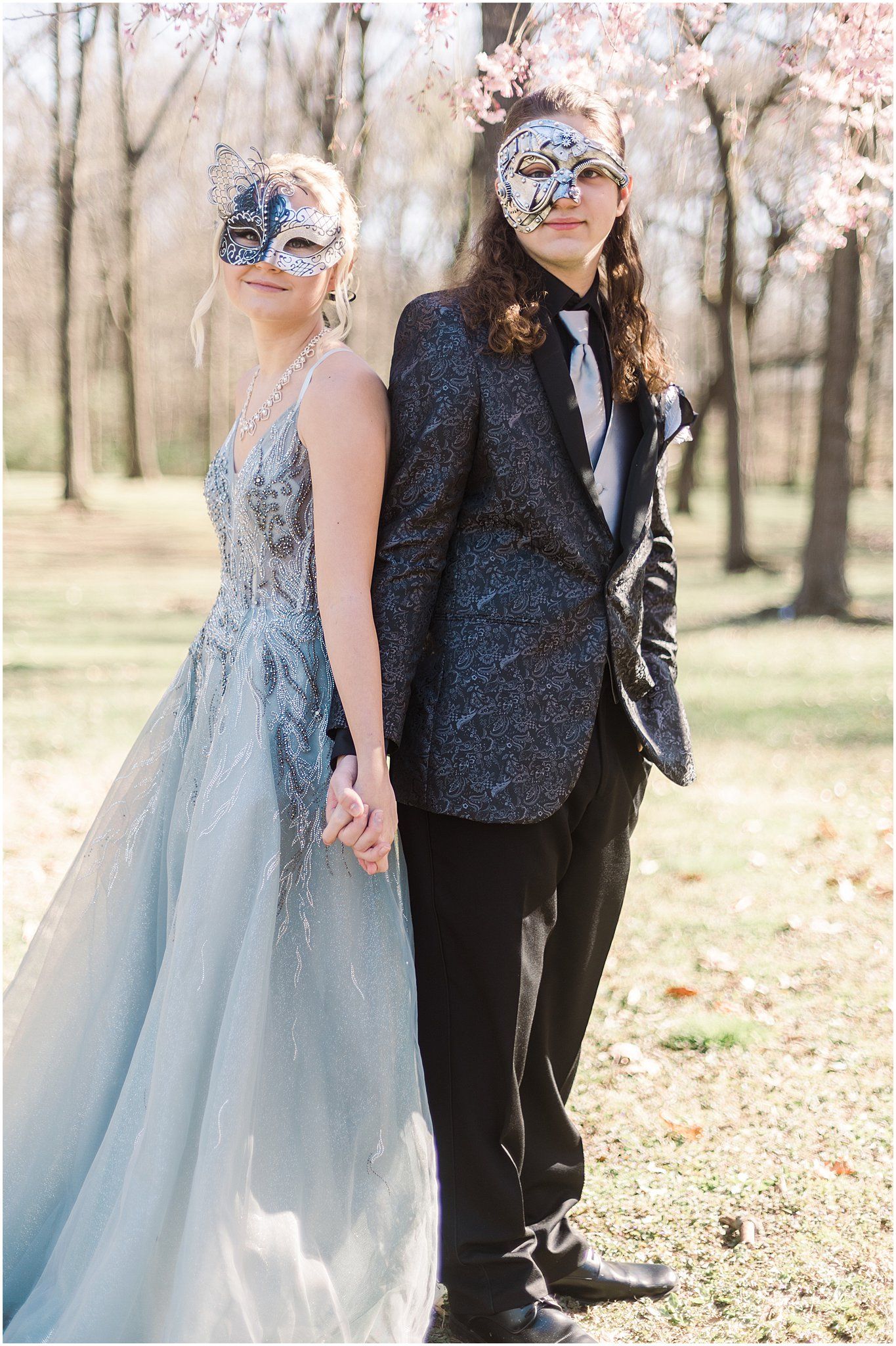 Elegant couple in masquerade attire pose against cherry blossom background.