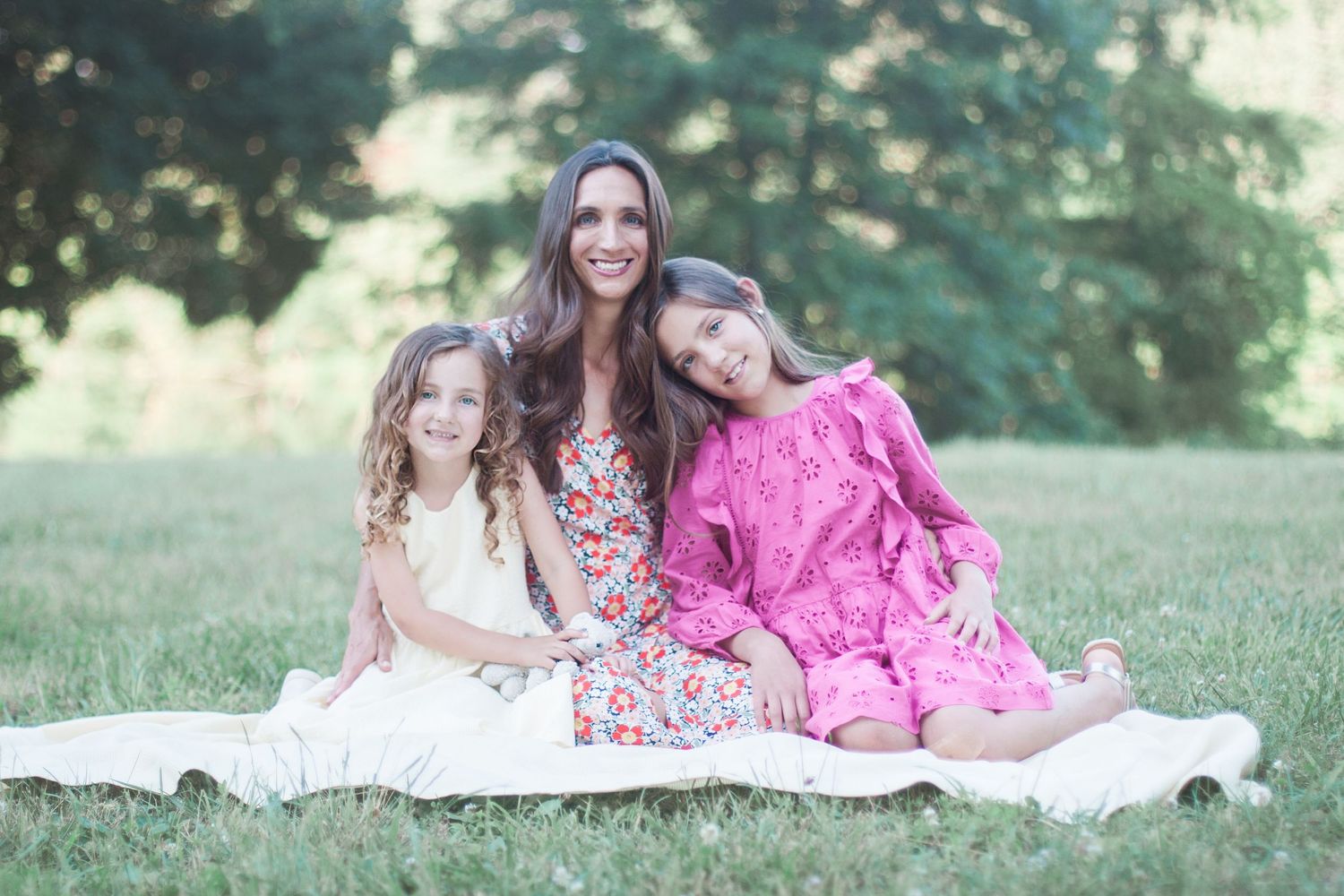 Outdoor family photo on blanket showing three people in summer dresses against green background.