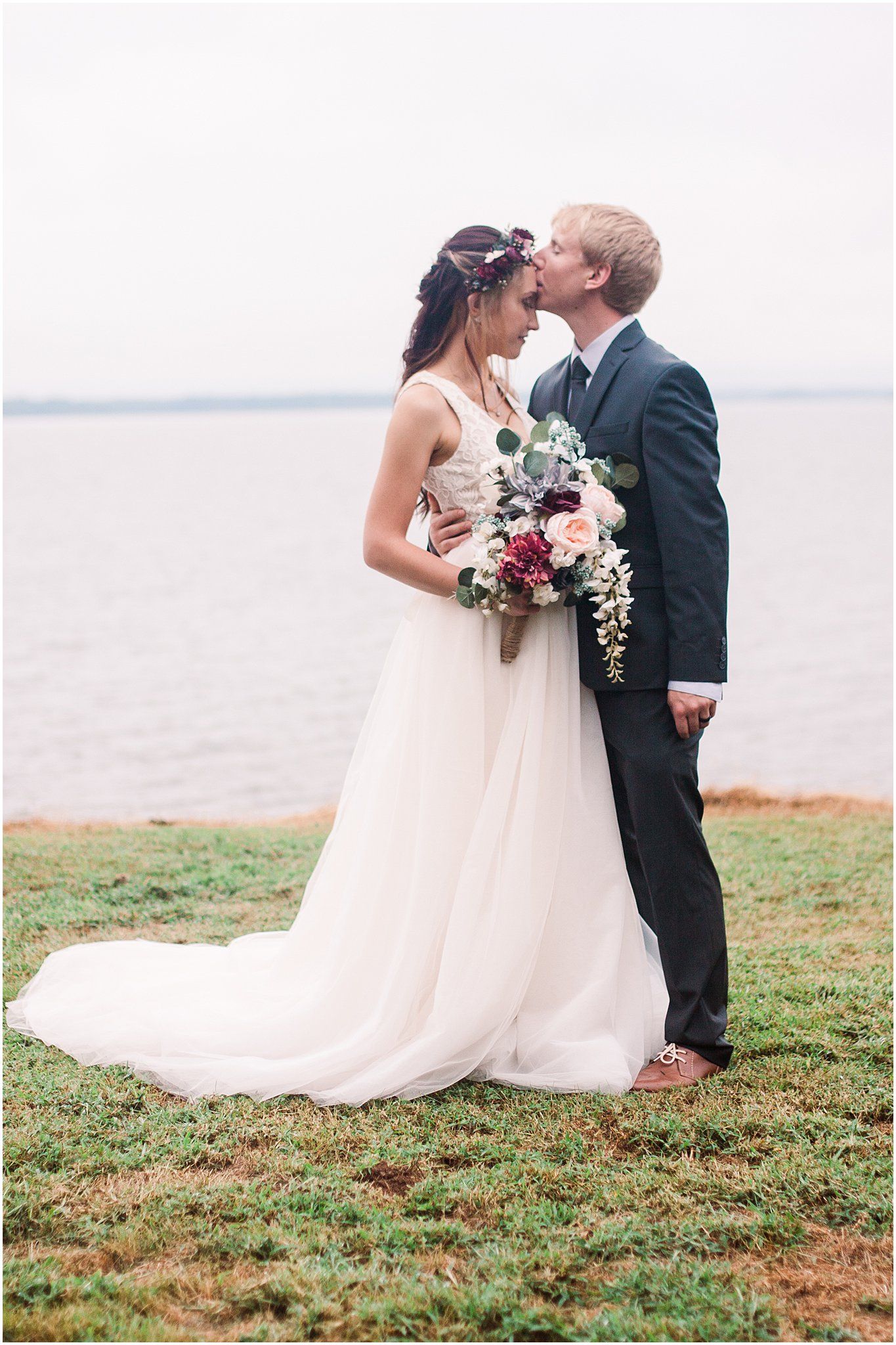 Romantic kiss between newlyweds on grassy waterfront with flowing white dress.