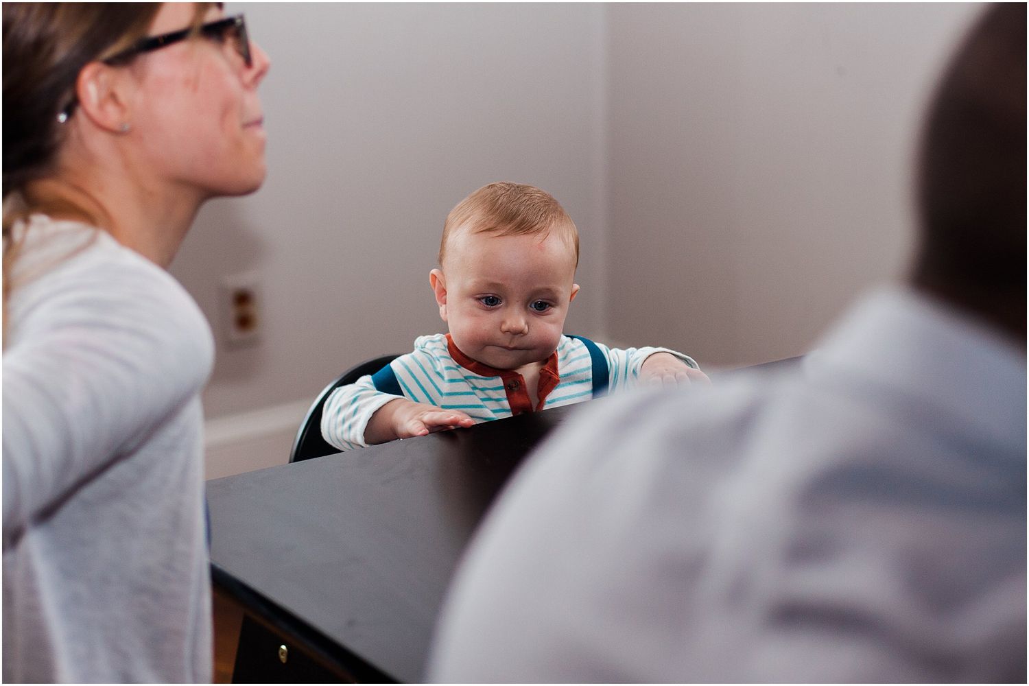 A baby peers over a dark table during a social gathering with adults.