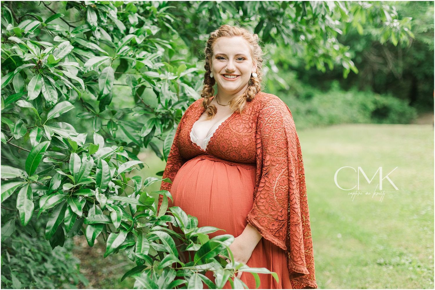 A maternity photo showing an expectant mother in a coral lace dress posing among green foliage.