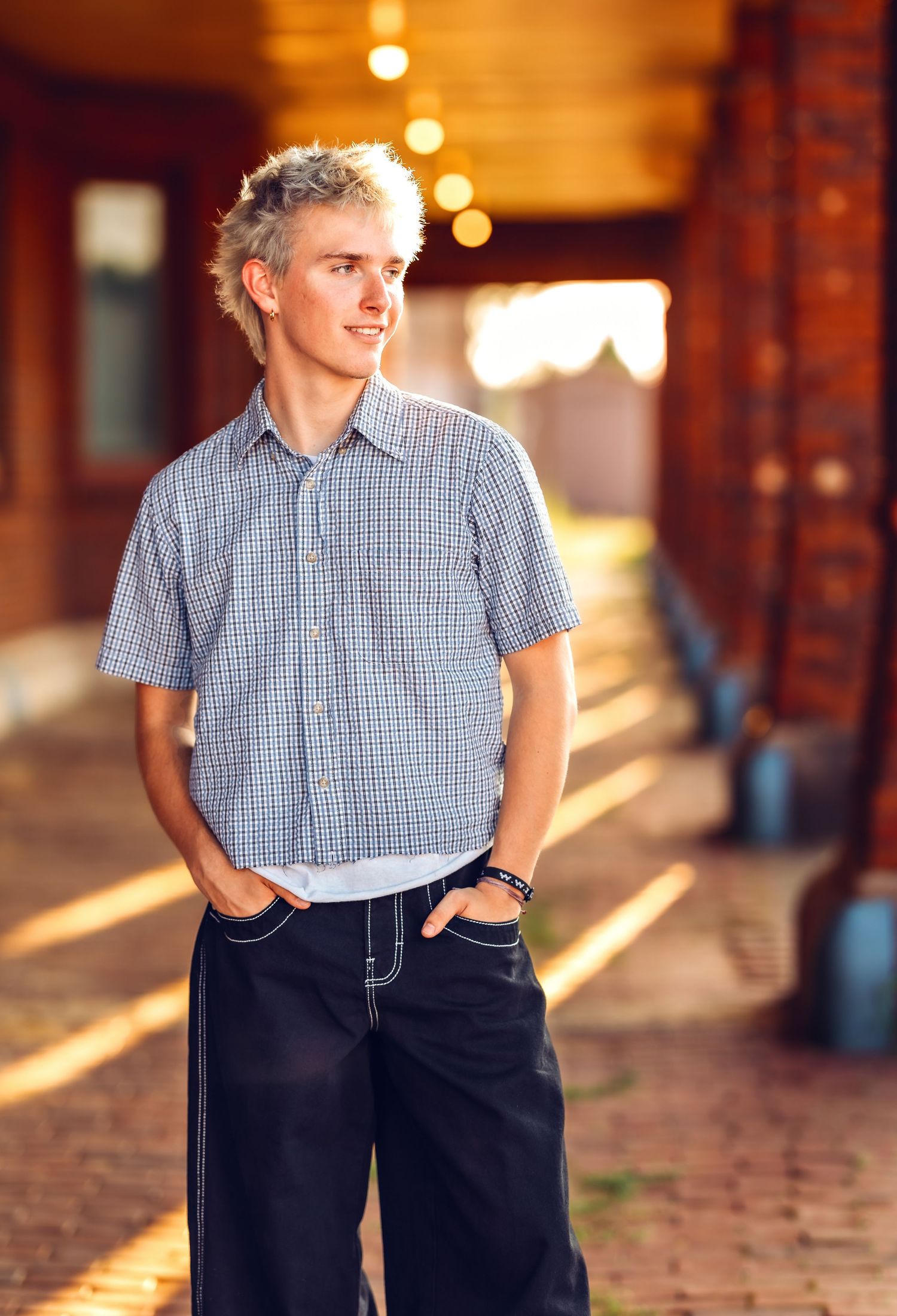 Figure in checkered shirt and dark pants poses in warm evening light against brick wall.