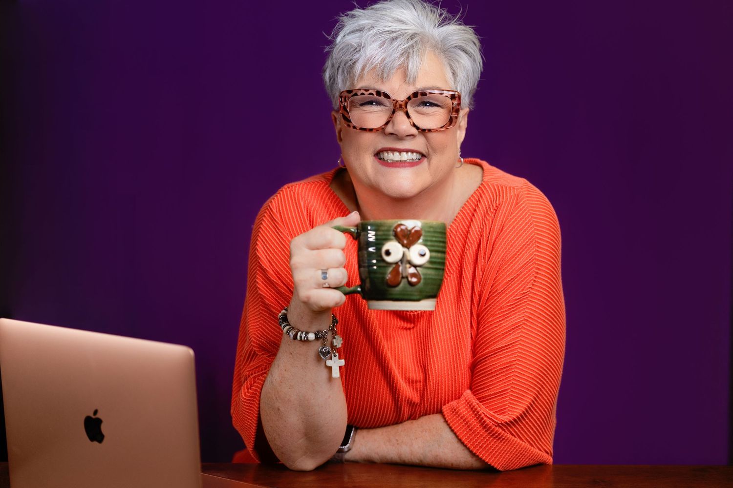 Smiling woman with short gray hair and glasses holds a festive mug beside a laptop against a purple background.