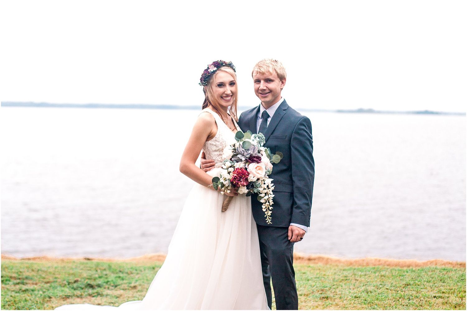 Newlywed couple poses with bouquet by waterfront on their wedding day.
