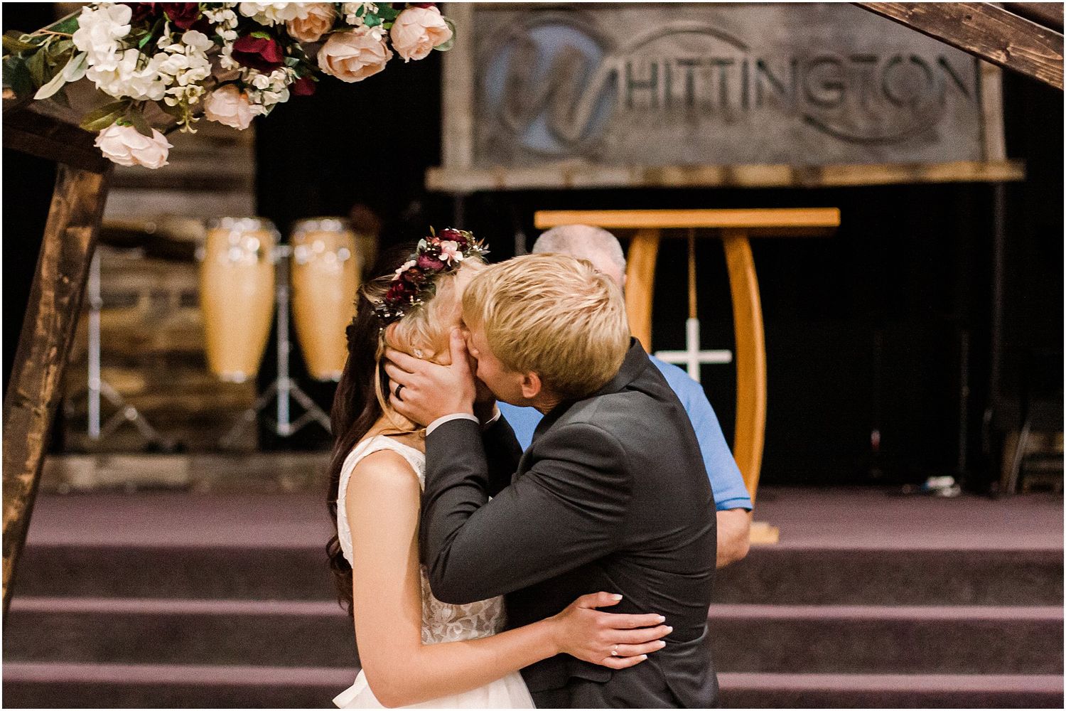 A couple shares their first kiss as newlyweds beneath a decorated wooden arch.