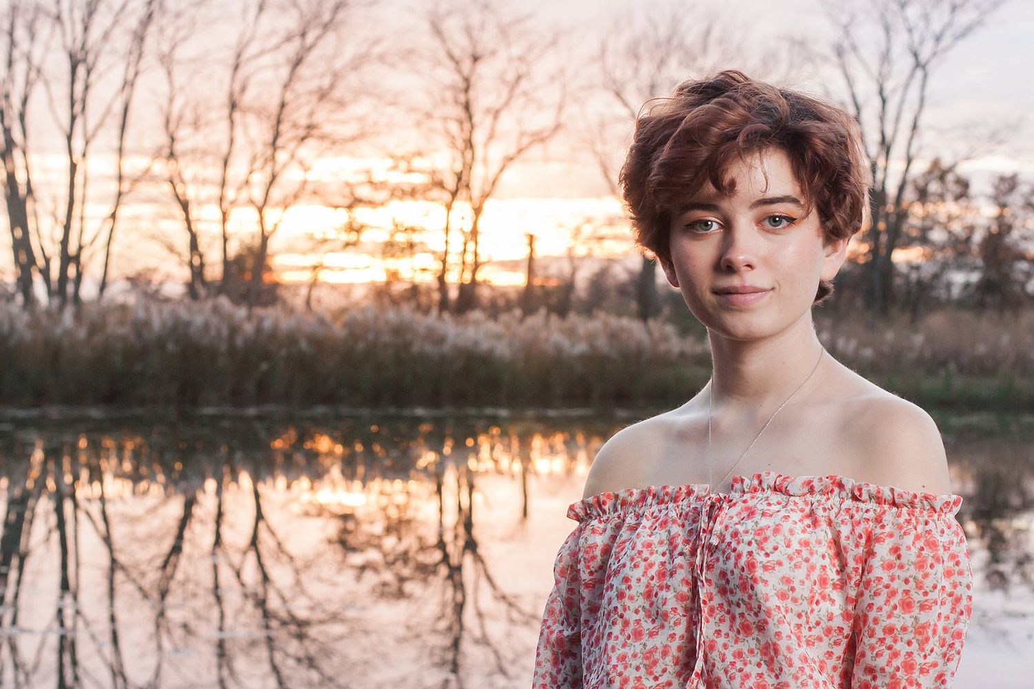 Portrait at sunset by reflective pond wearing floral off-shoulder dress.