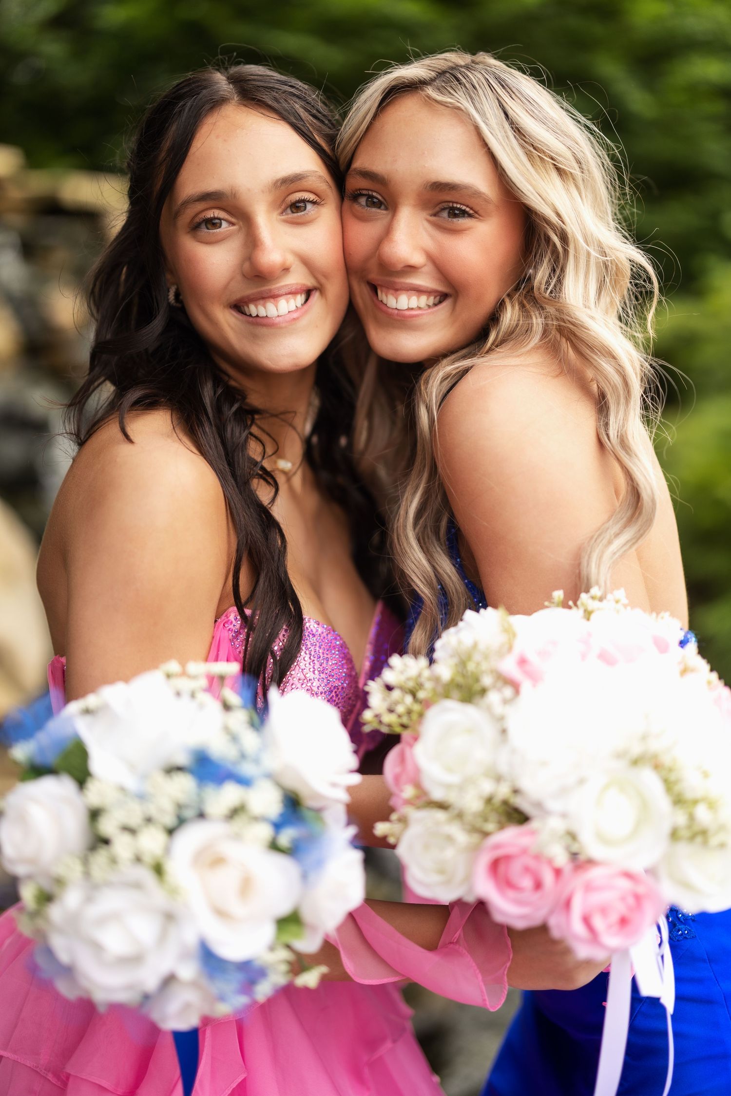 Two smiling teenage girls in pink and blue prom dresses holding floral bouquets pose together outdoors.