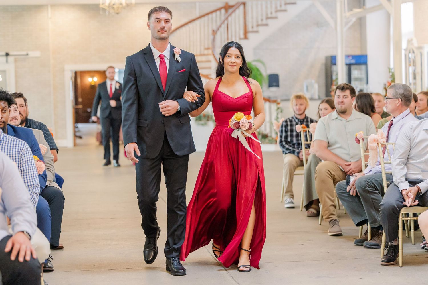 Wedding party members walk down the aisle in formal attire with guests seated on either side.