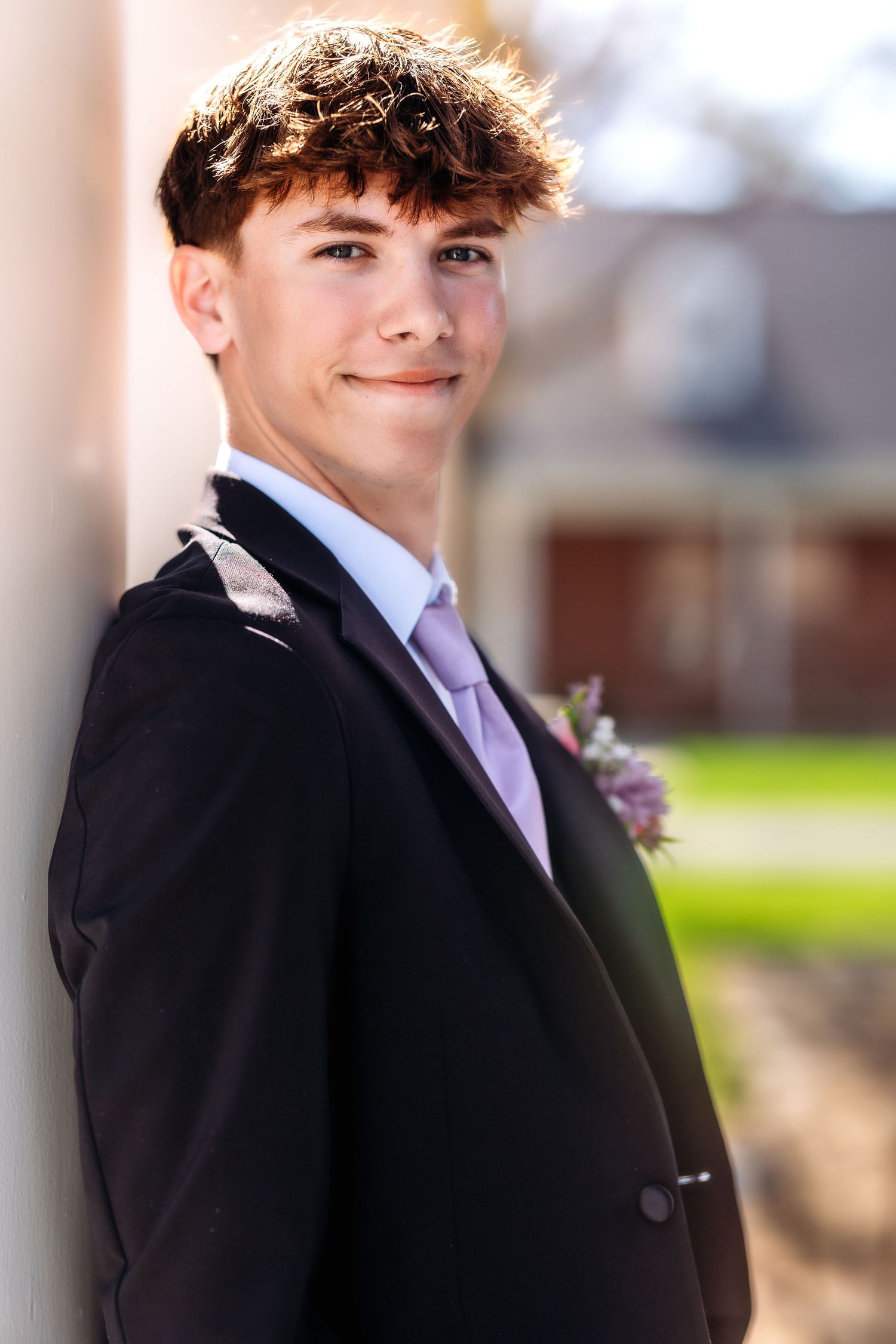 A person in a black suit with a purple tie and boutonniere poses outdoors in a formal portrait setting.