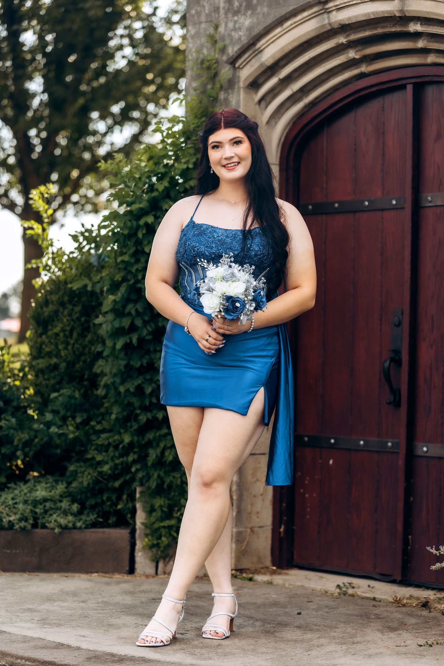 Young woman in blue mini dress holding white bouquet poses by ornate wooden doors with greenery backdrop.