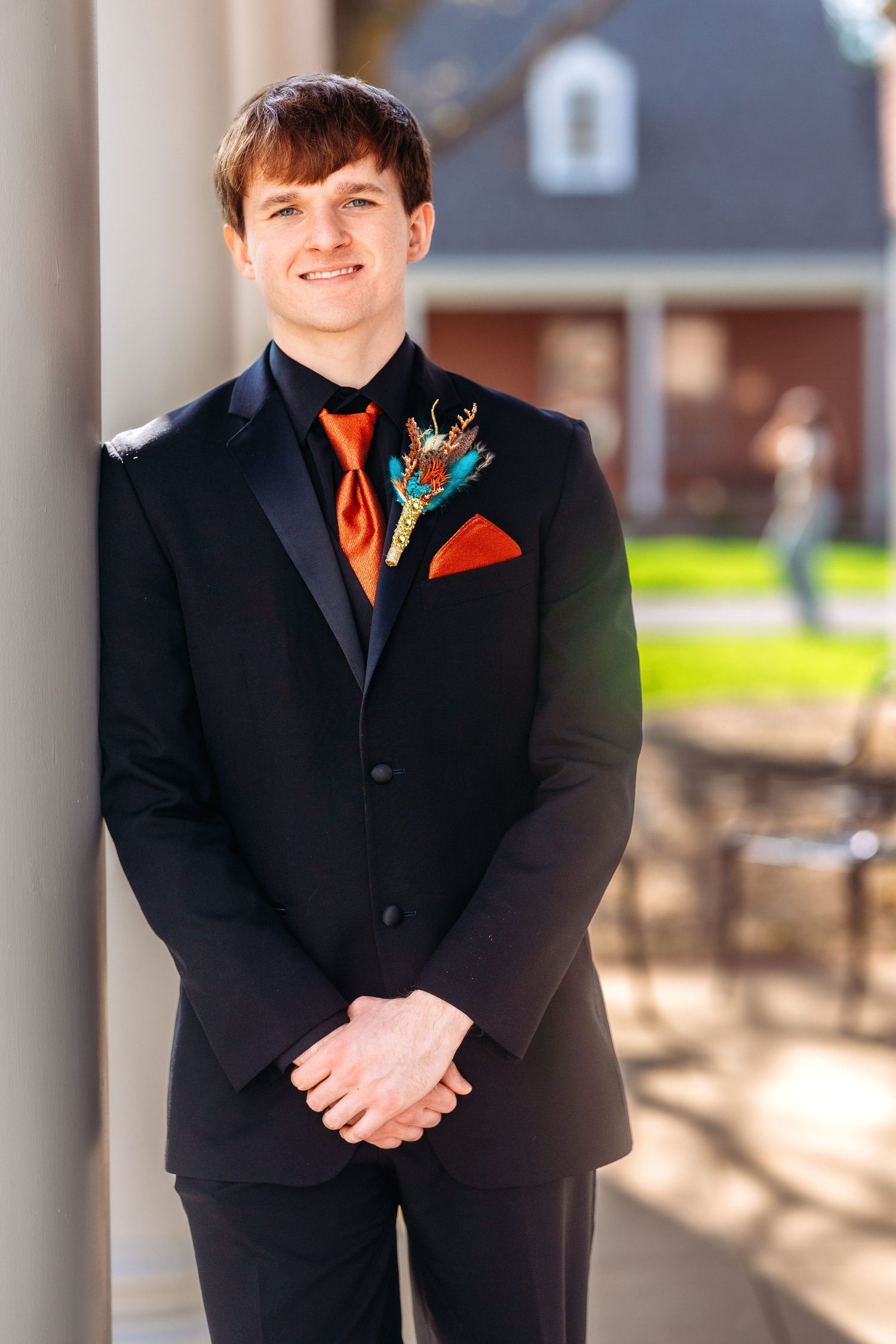 A person wearing a black suit with orange boutonniere poses in front of stone columns on a sunny day.
