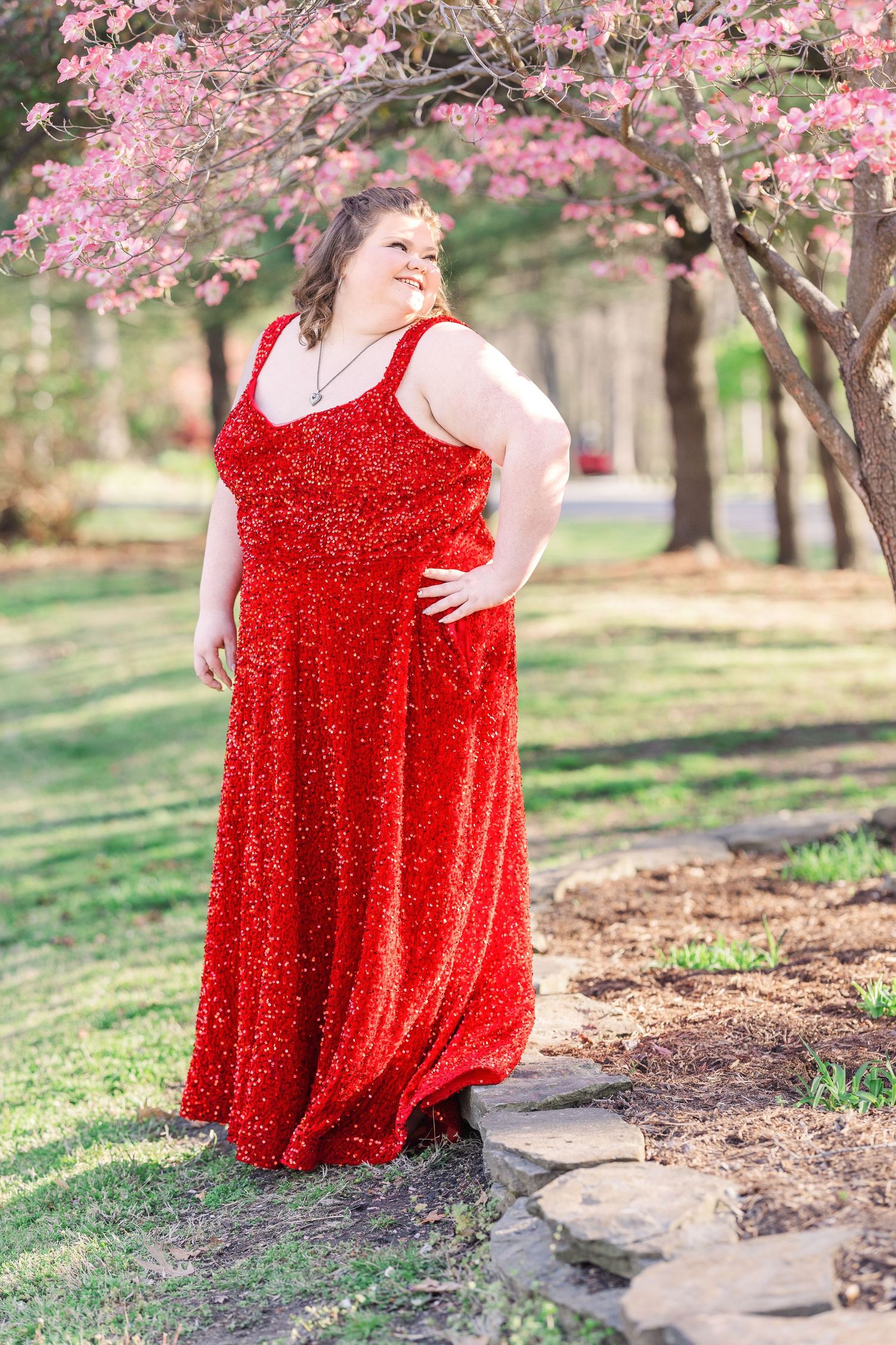 Person in a sparkly red formal gown poses under blooming cherry blossom trees in a park setting.