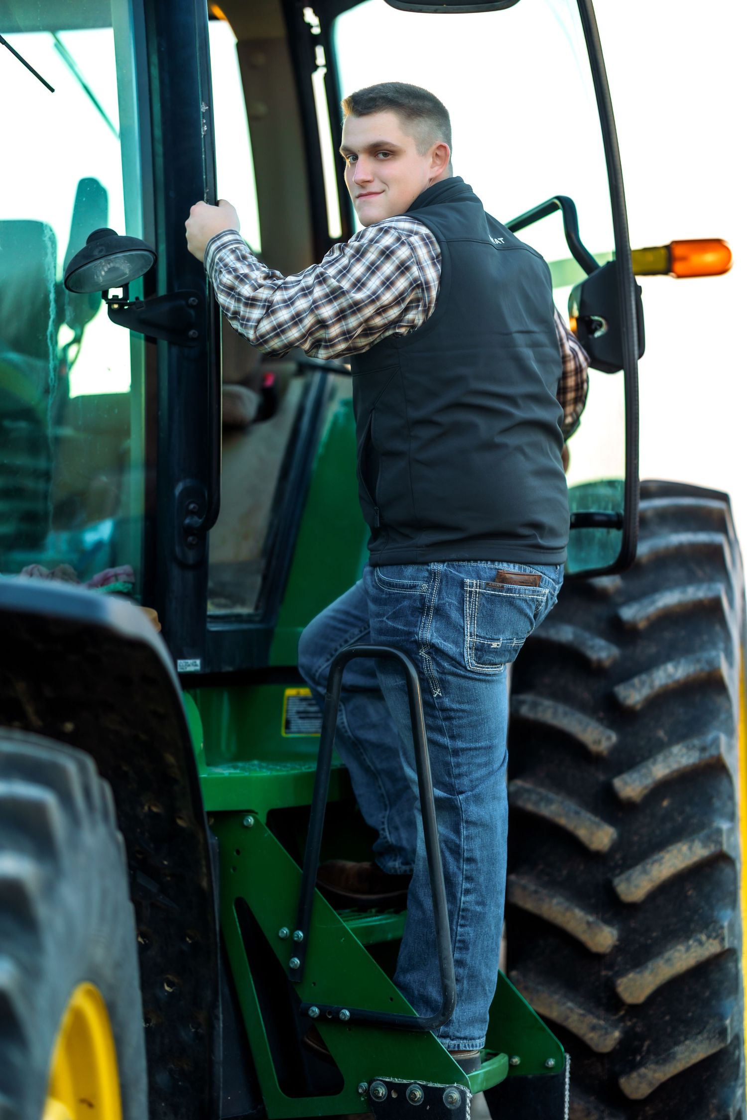 Young farmer climbing into green tractor cab wearing plaid shirt and vest.