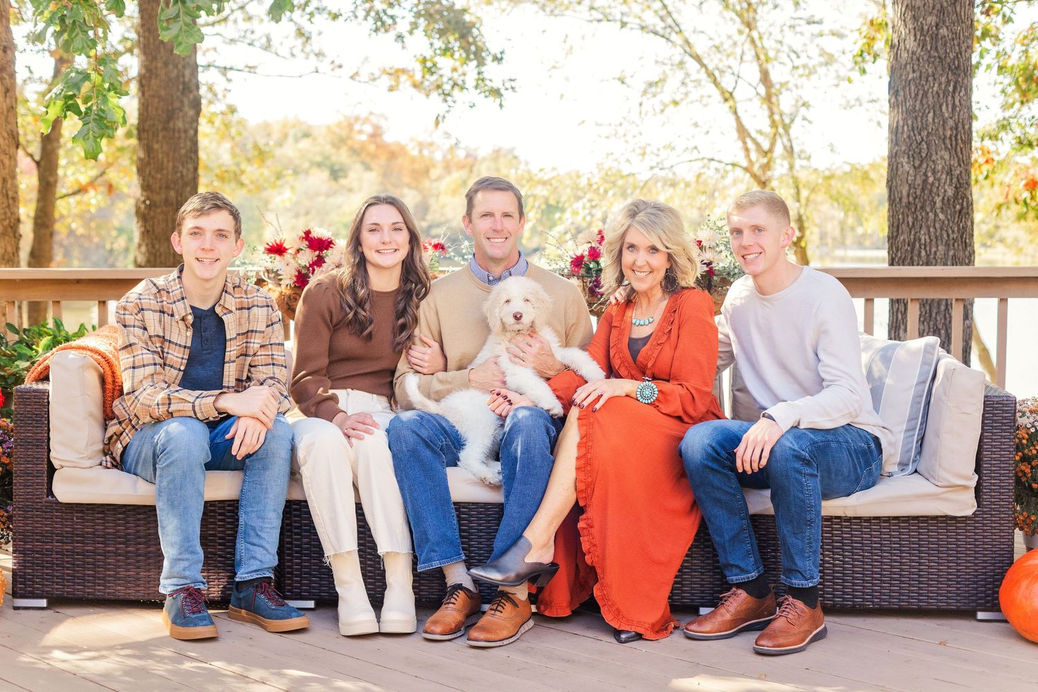 A family sits together on outdoor patio furniture with their small white dog during an autumn photo session.