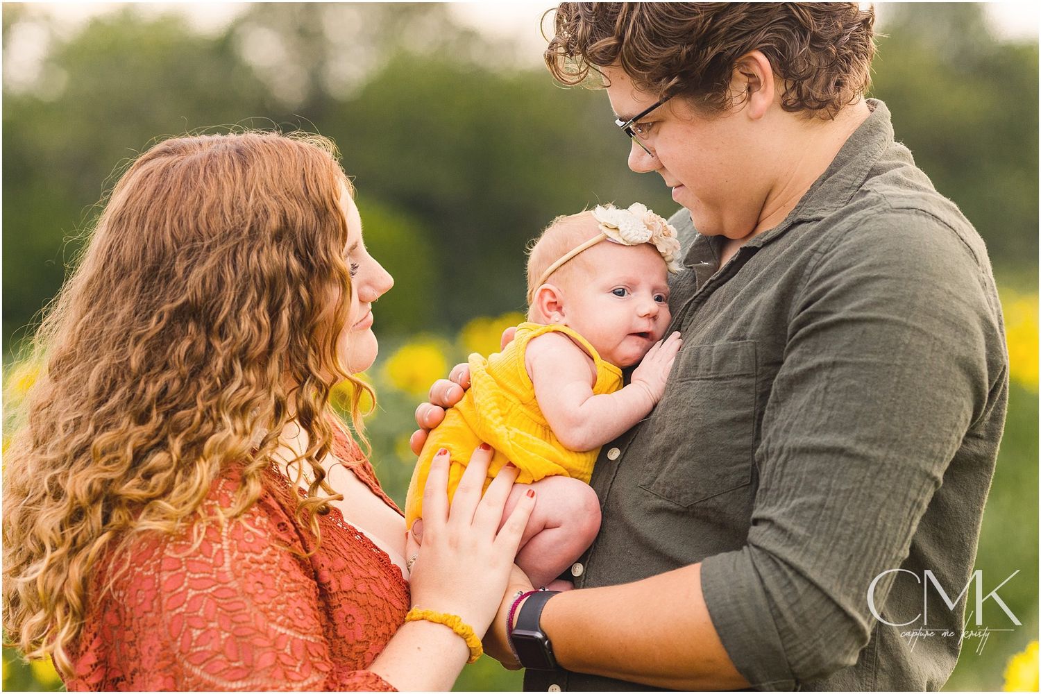 Parents share a tender moment with their infant wearing yellow in a sunny outdoor setting.