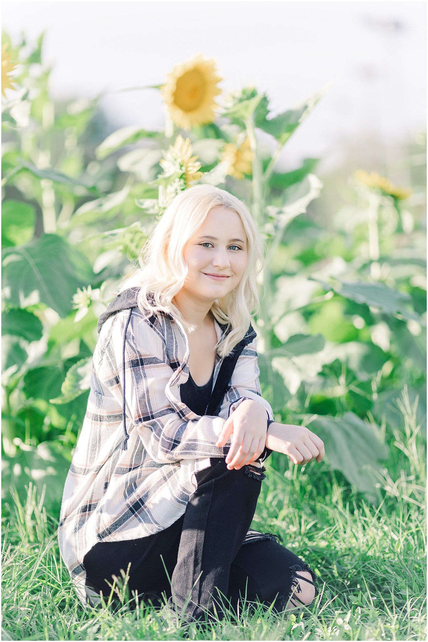 Senior girl with blonde hair kneels in front of sunflowers with sun shining behind her