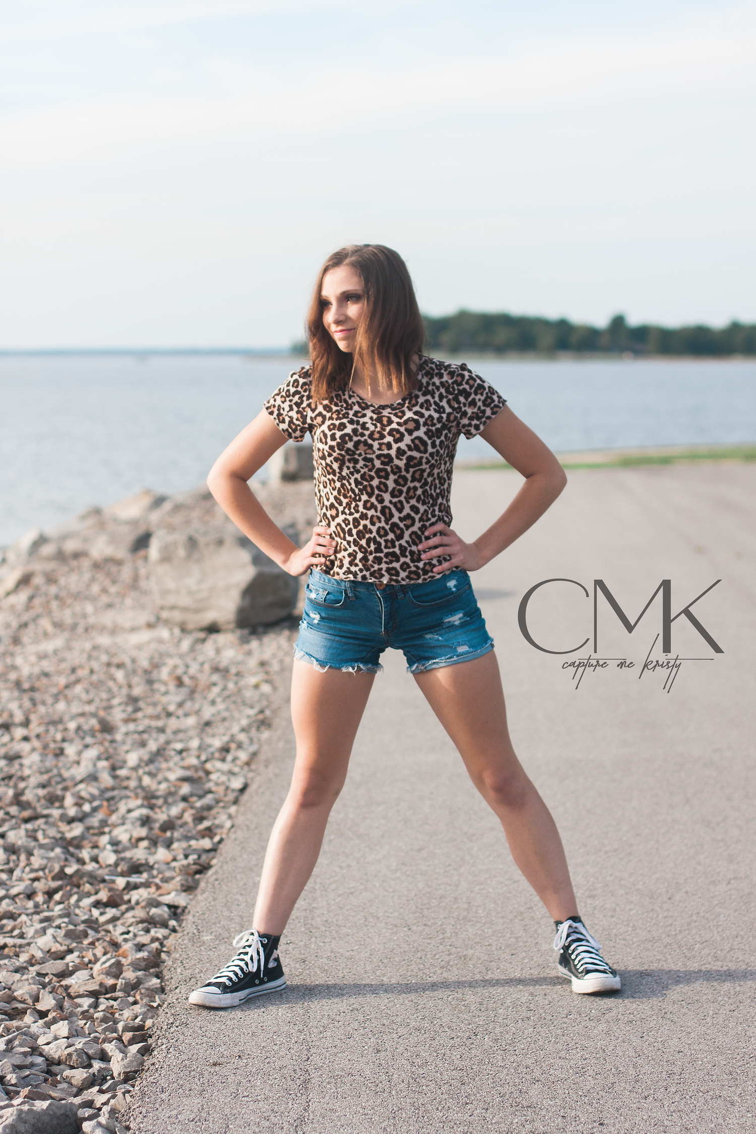 Dynamic pose in leopard print top and denim shorts against waterfront background.
