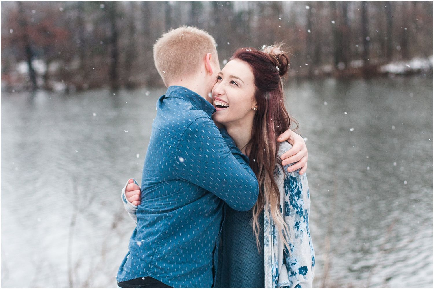 A couple embraces and laughs together during a snowy outdoor photo session near a lake.