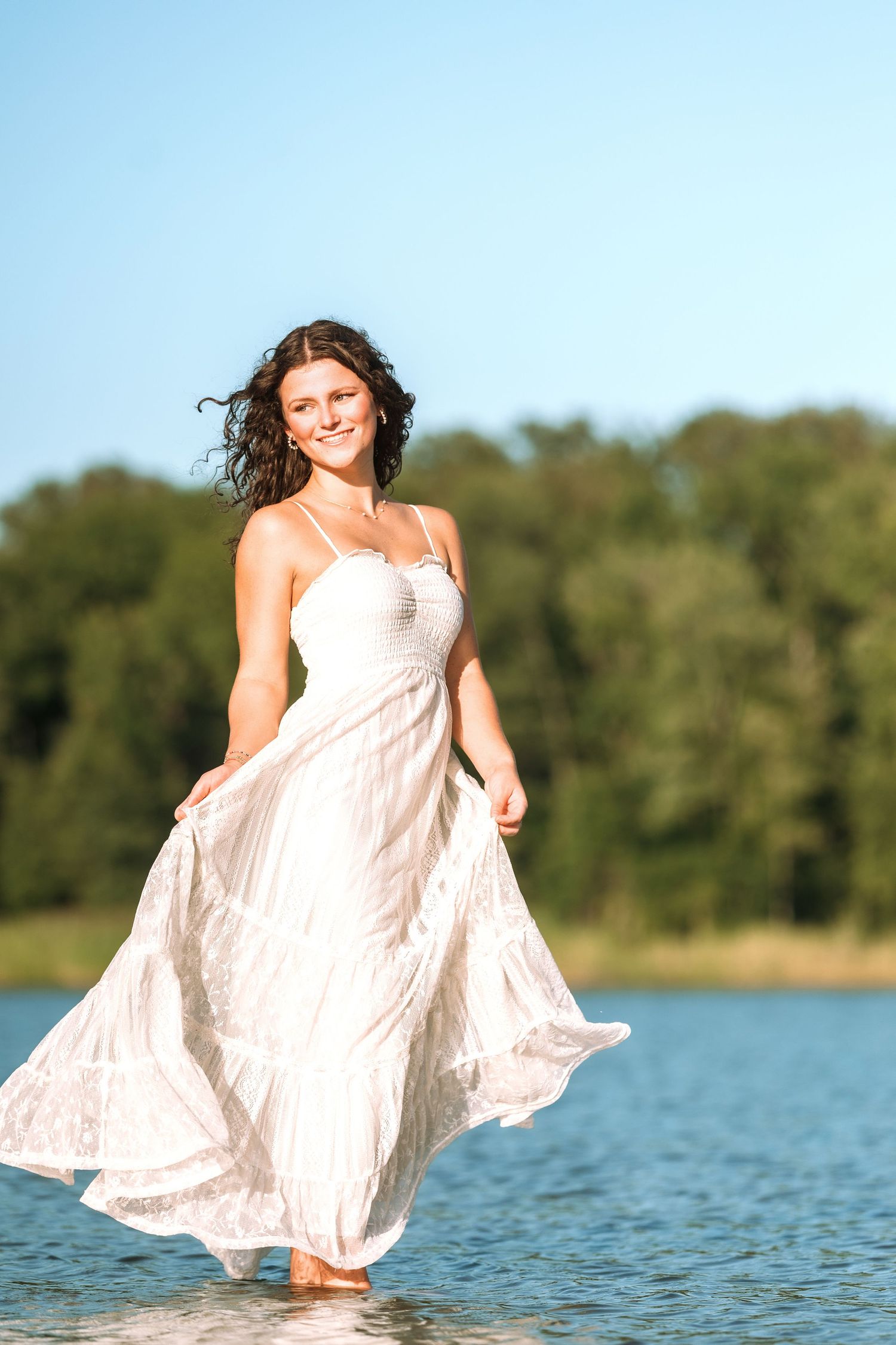 A smiling woman in a flowing white dress wades through water with trees and blue sky in the background.