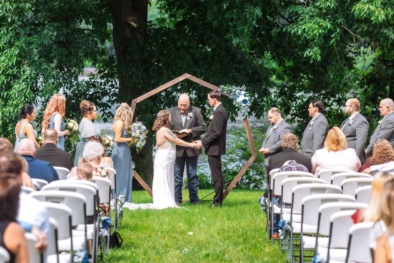 Outdoor wedding ceremony with white chairs and wooden arch surrounded by lush greenery.
