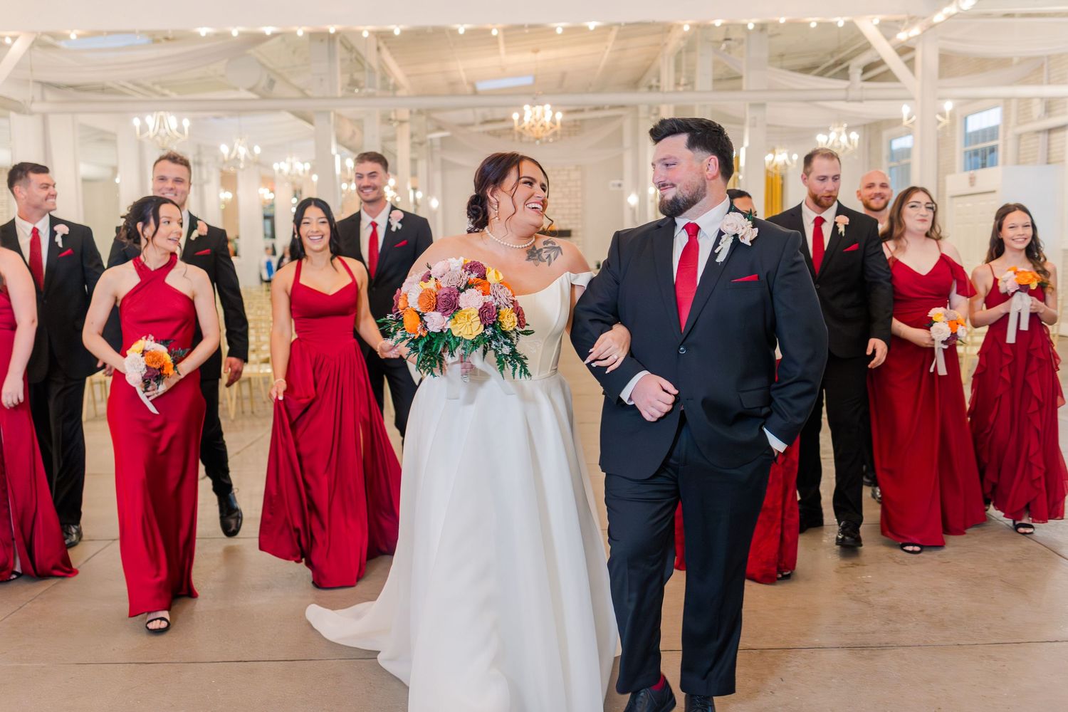 Wedding party members walk in pairs down an elegant white indoor venue with red accents.
