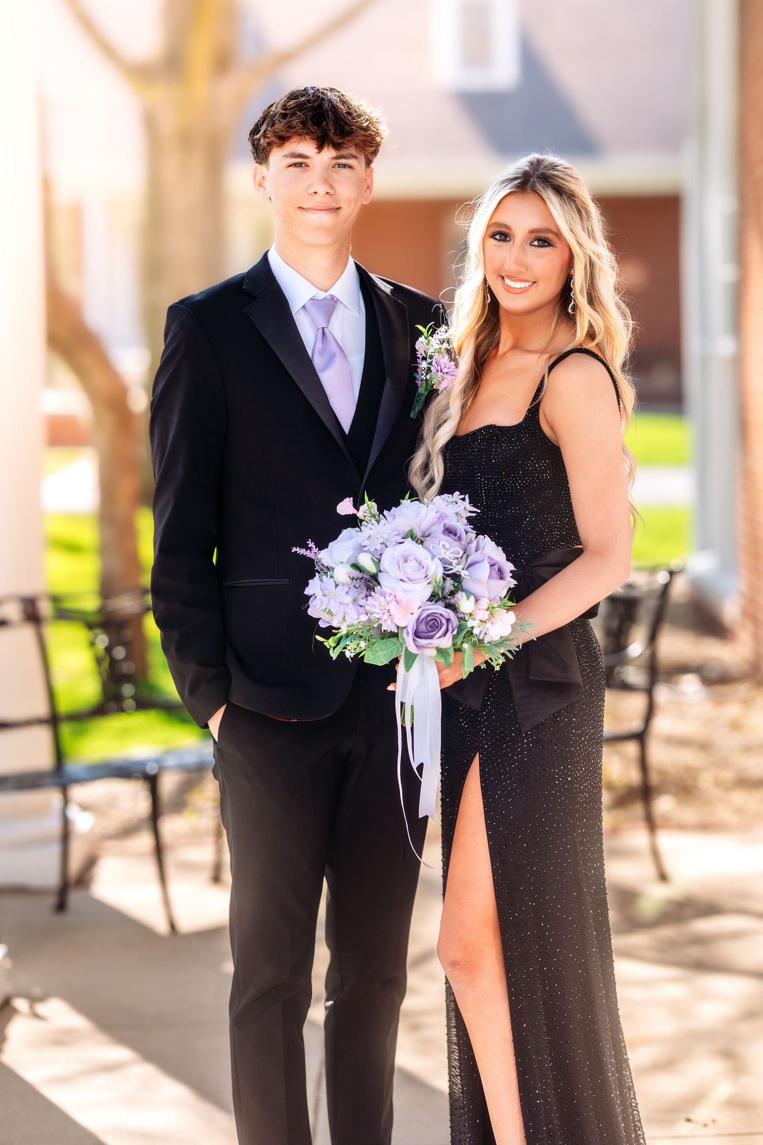 A couple poses for a formal photo, with the woman holding a purple and white bouquet and wearing a black dress.
