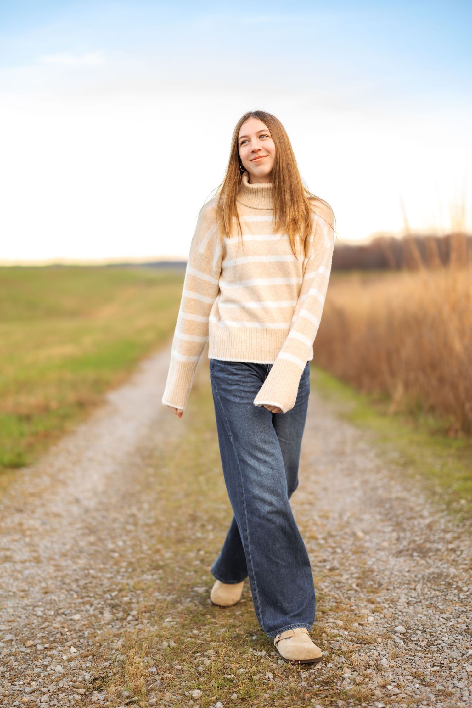 Person in cream sweater and jeans walking on a gravel path during golden hour.