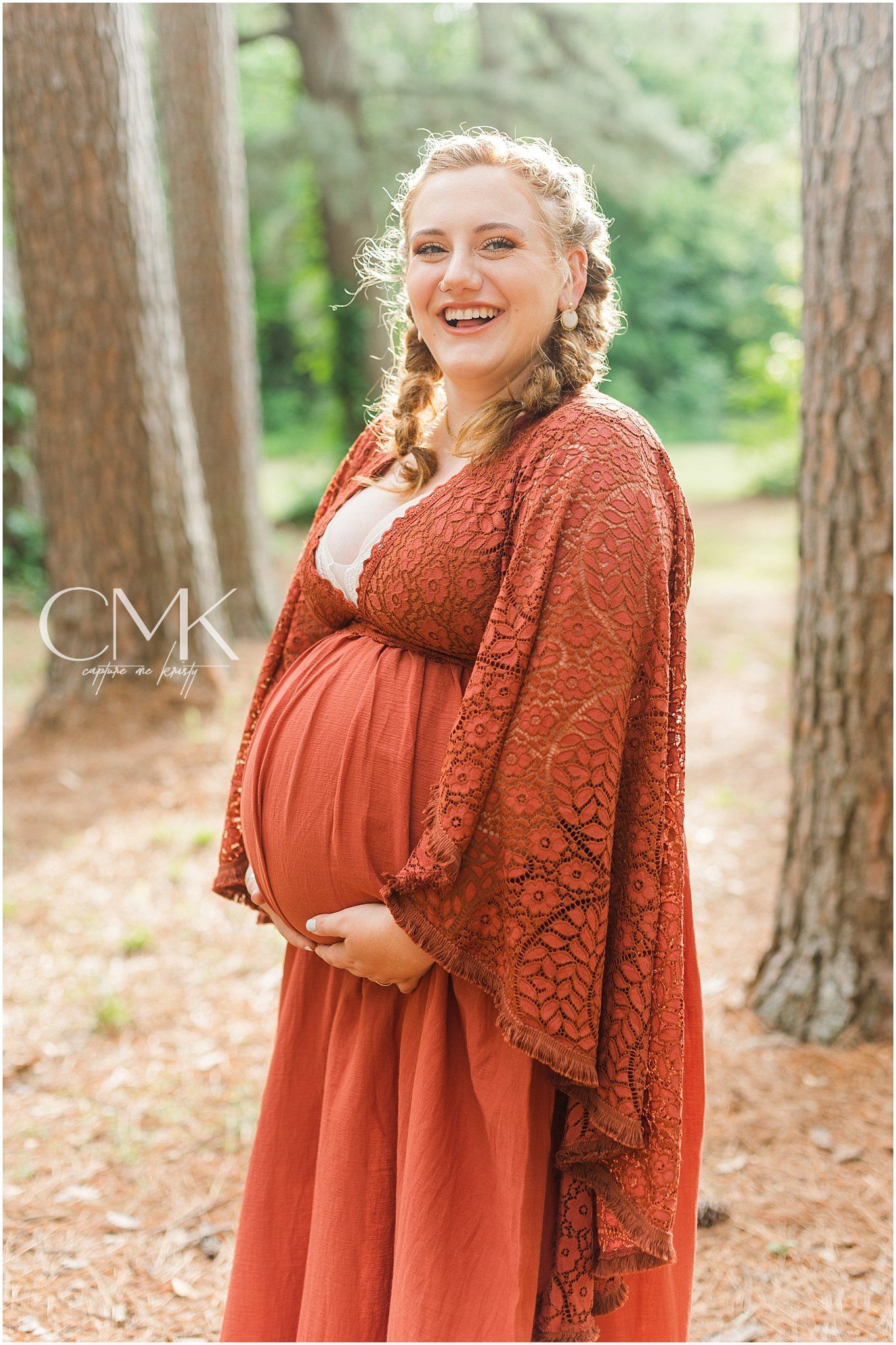 Expectant mother in rust-colored lace dress smiles radiantly in pine forest setting.